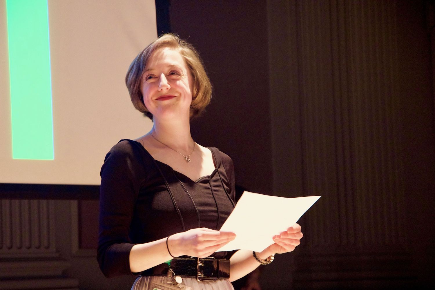 Female-presenting person smiling, head turned right, holding a piece of paper with both hands.