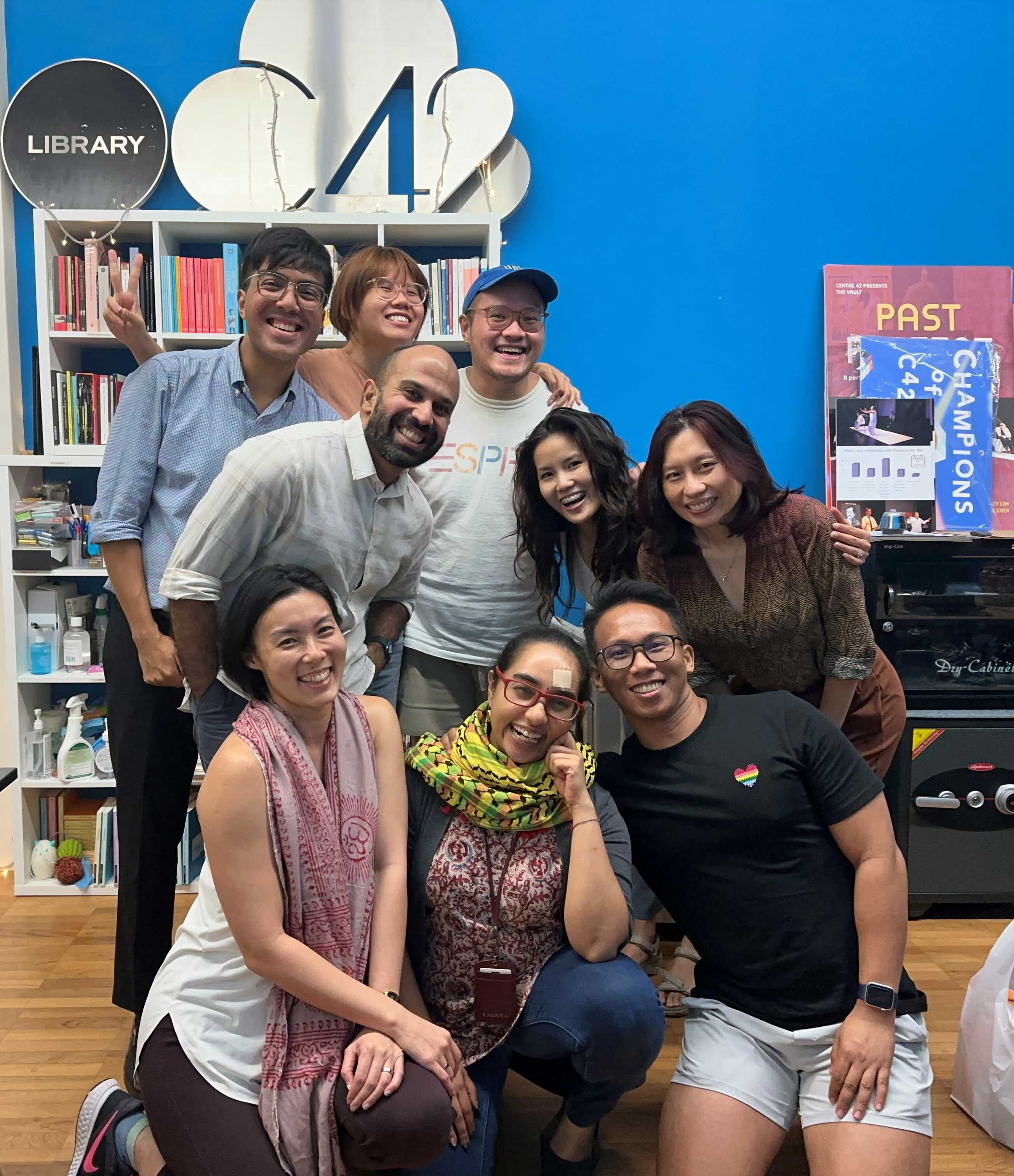 A group of 9 people posed in front of a shelf of books. On top of the shelf sits a black round signage with the text LIBRARY and C42.RY