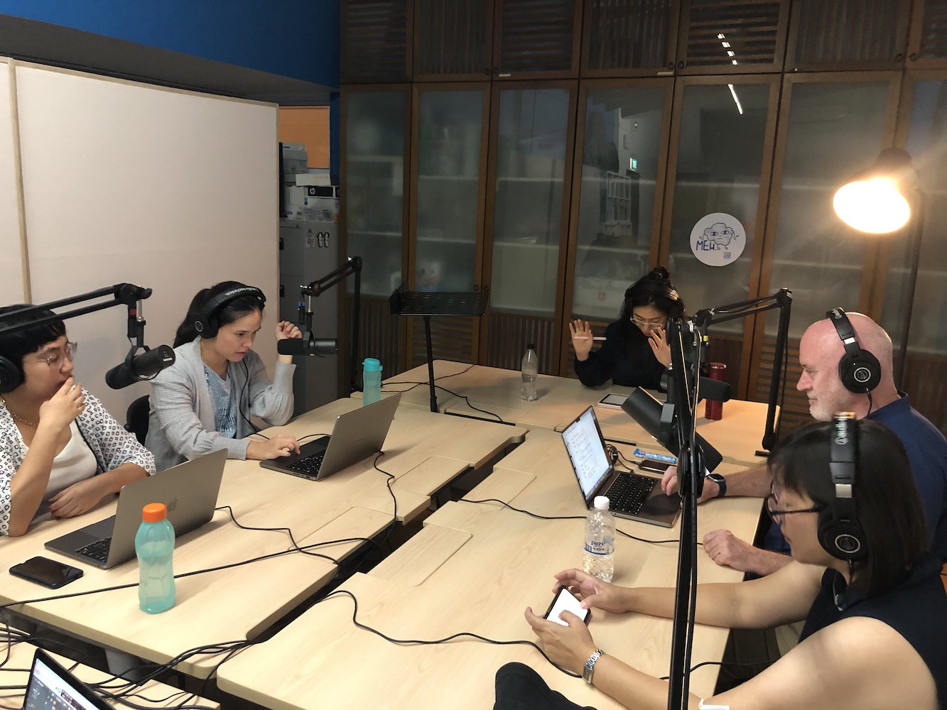 Five persons wearing headphones seated around a table, facing their laptops and cellphones.