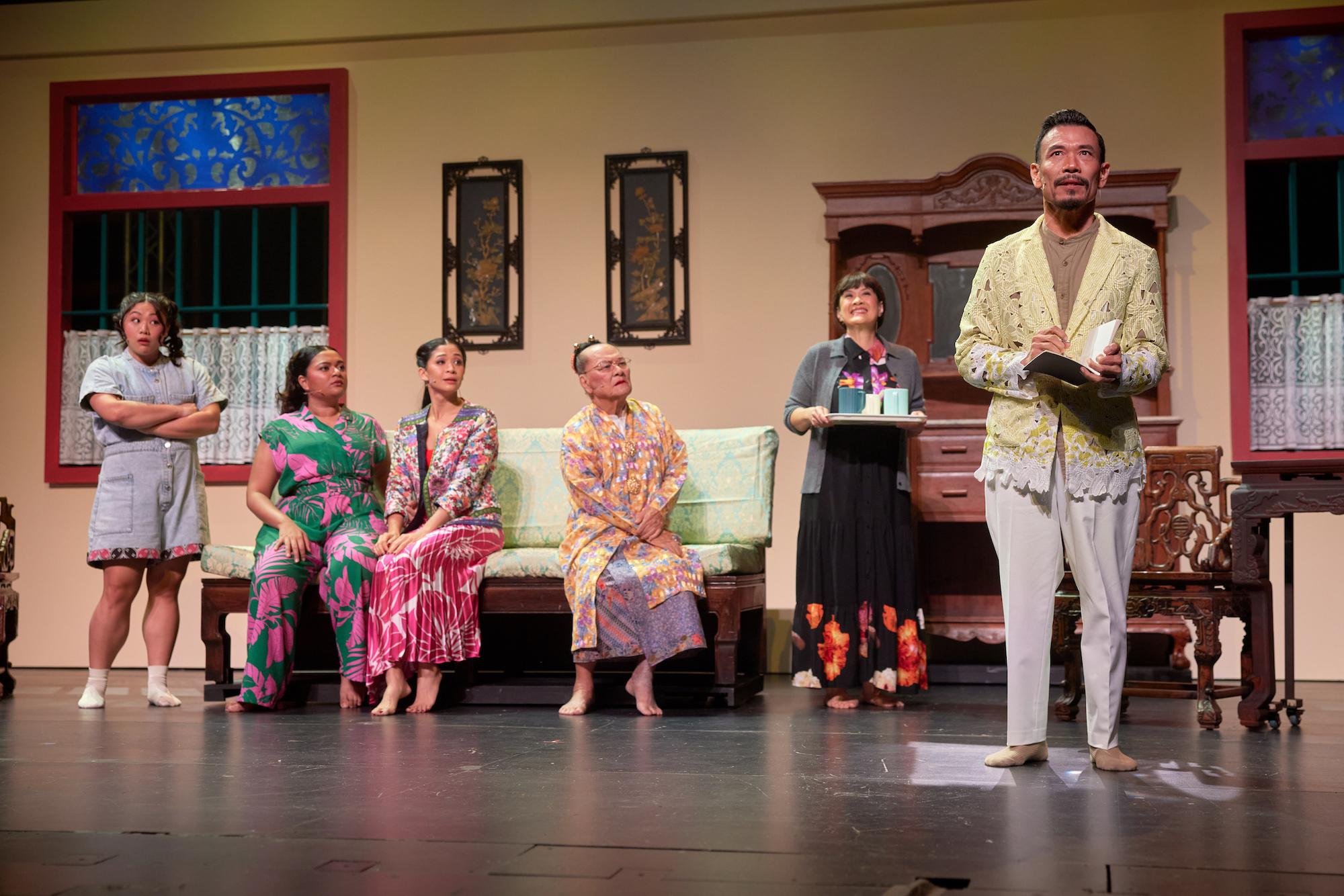 A male-presenting person standing on a stage holding a notebook. Behind him are five people sitting on a sofa, watching him.