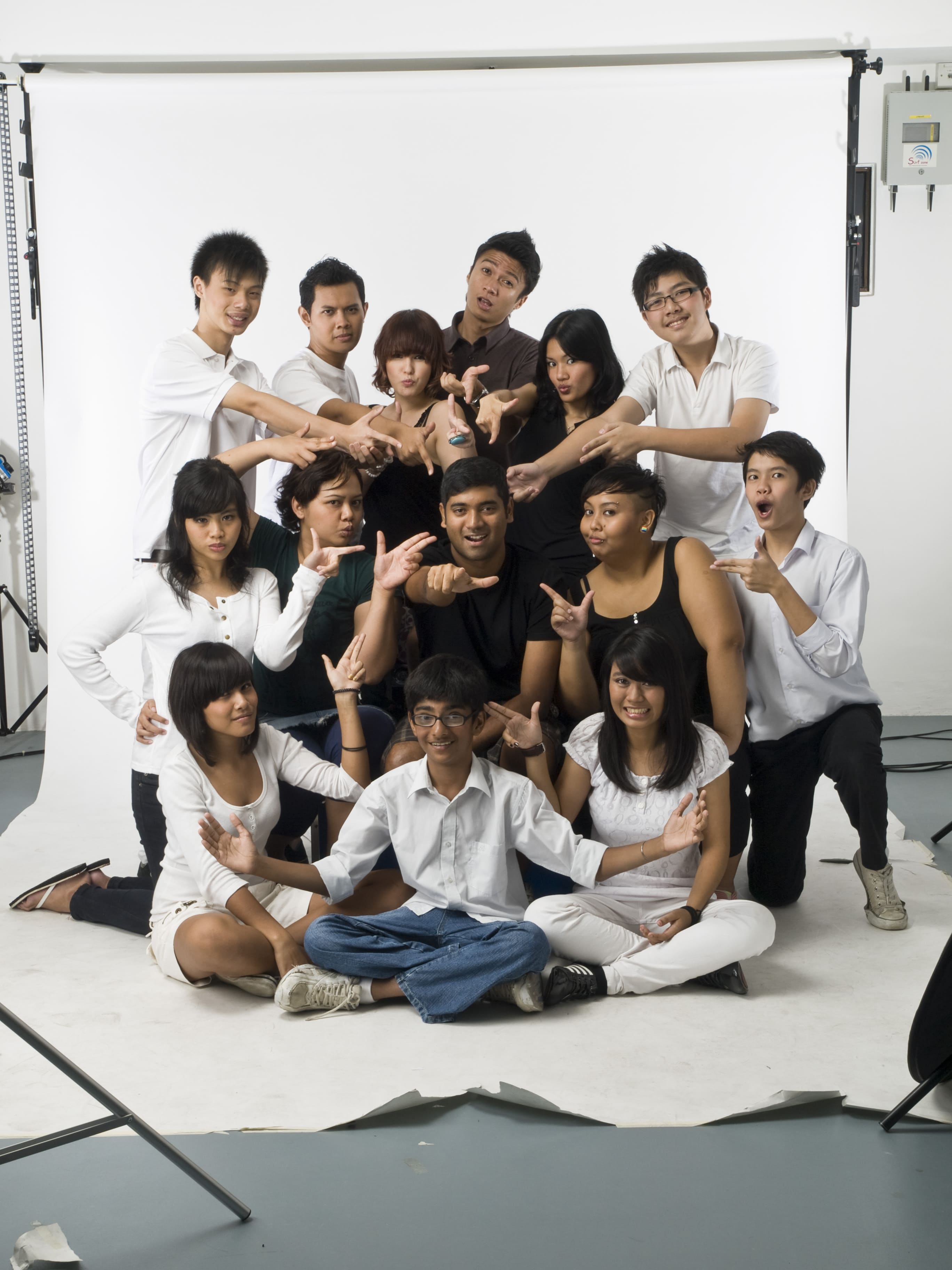 The photograph features fourteen persons arranged in a cluster against a white backdrop, in what appears to be a photo studio. The person in the center is pointing a finger gun at the camera, while the rest are pointing finger guns at him.