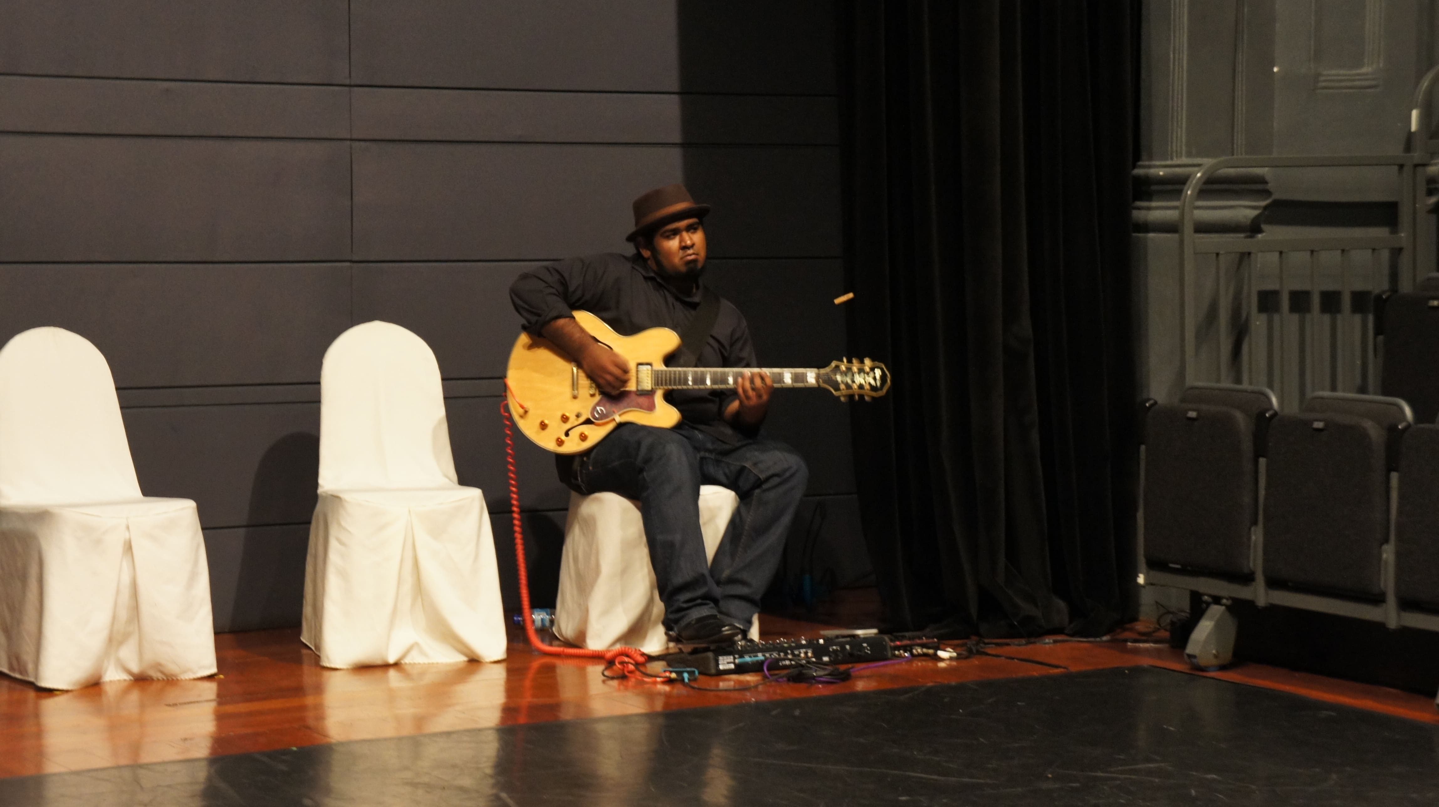 The photograph features a male presenting person wearing black clothing and a black hat sitting on a white chair, playing a guitar. There are two empty white chairs to his left.