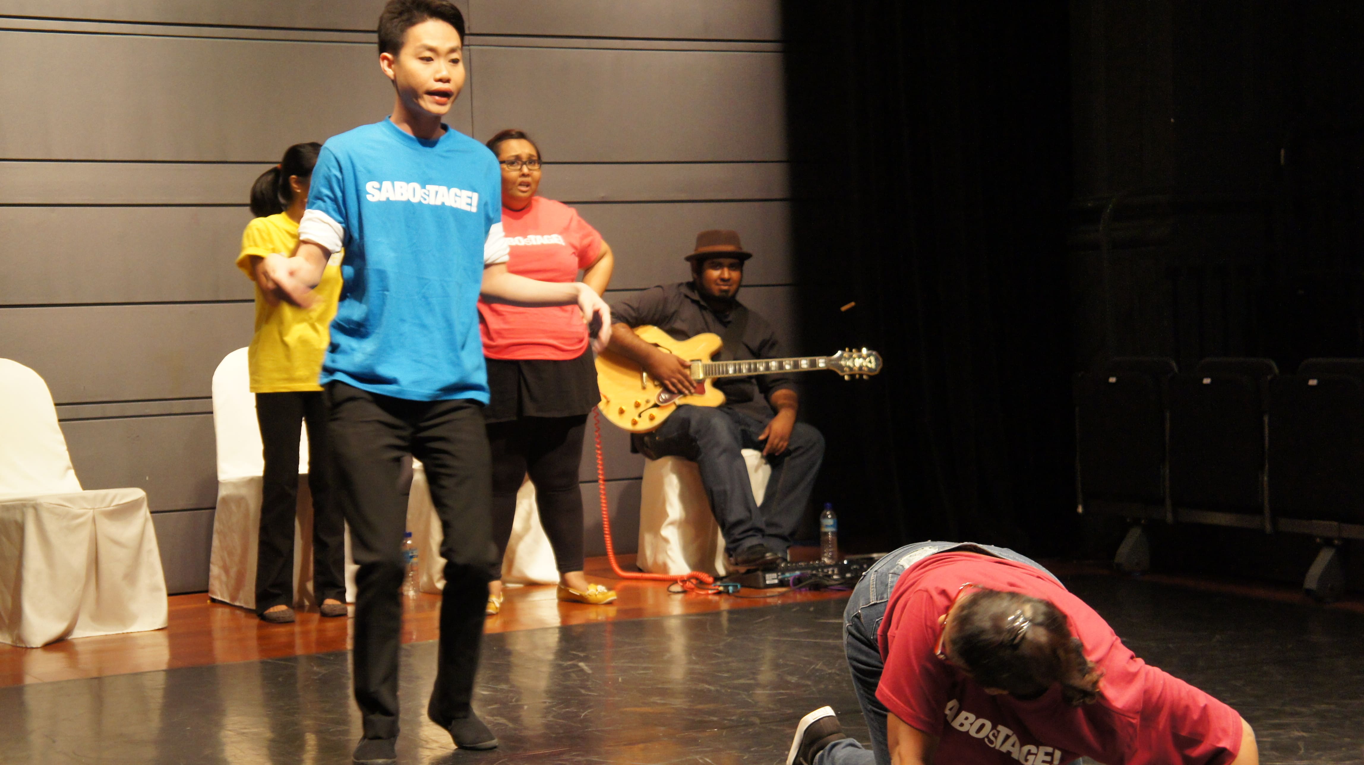 The photograph features a male presenting person wearing a blue shirt bearing the words SABOsTAGE, standing behind a female presenting person wearing a pink shirt who is prostrating towards the audience, her head turned to face the male presenting person.