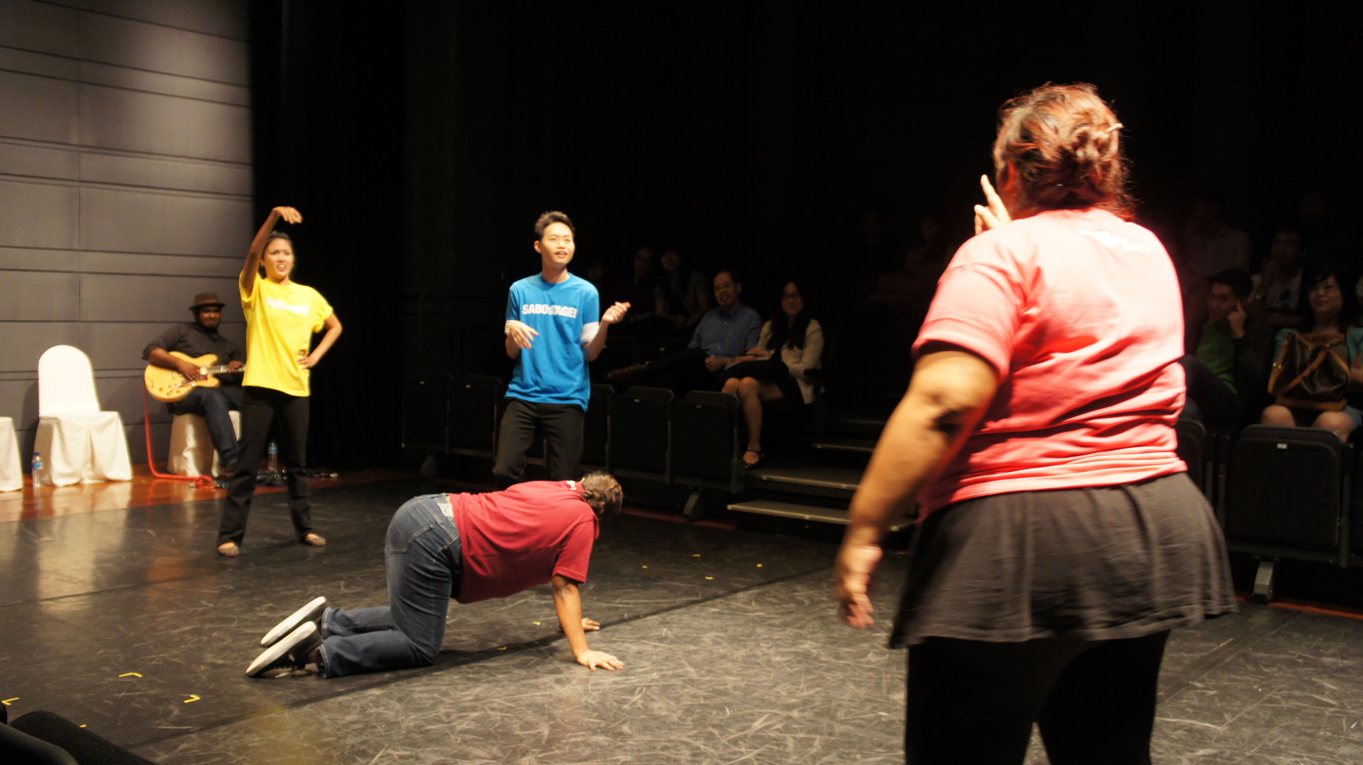 The photograph features a female presenting person wearing a pink shirt on all fours, with two persons wearing yellow and blue to one side of her, speaking to another person wearing pink on the other side.