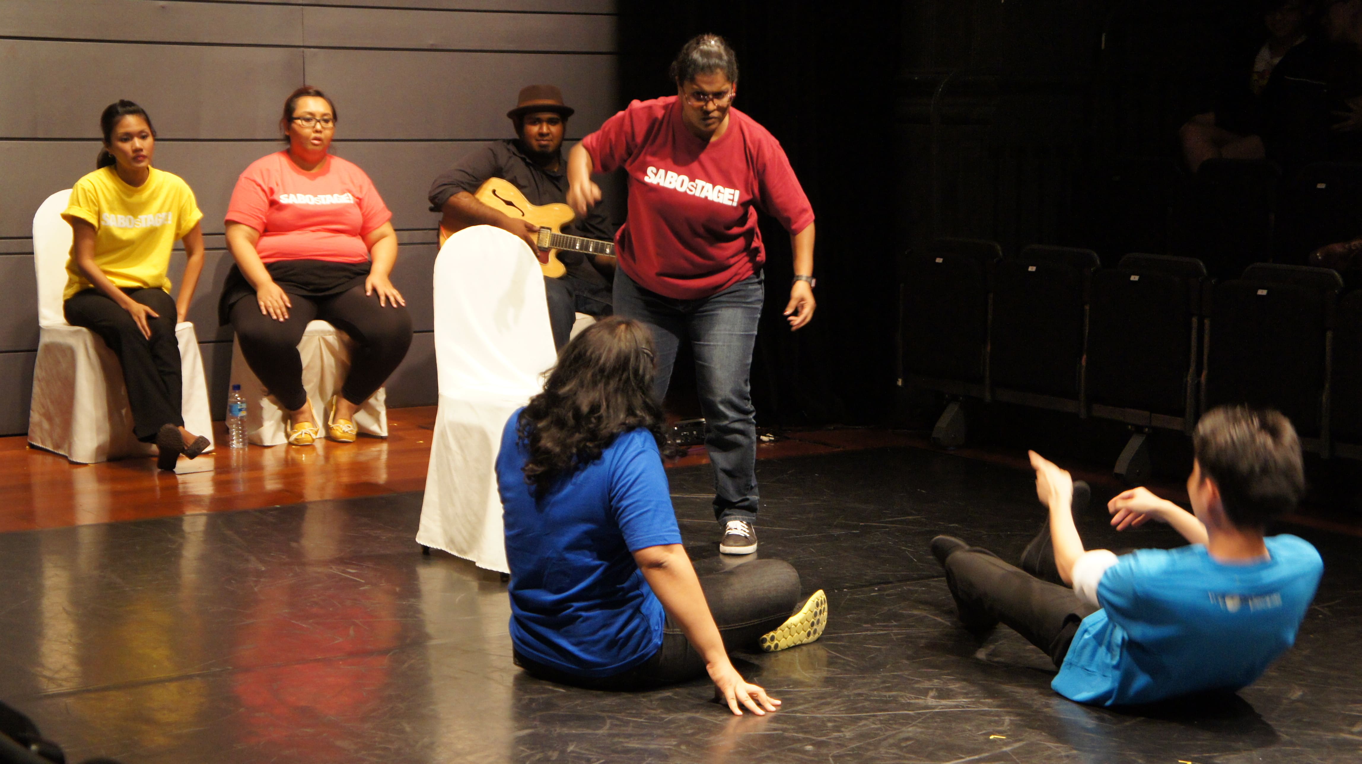 The photograph features a female presenting person wearing a red shirt raising her right hand while glaring at two persons in front of her, one seated in a cross-legged position while the other is leaning back with legs outstretched. The rest of the performers are seated at the back on white chairs.