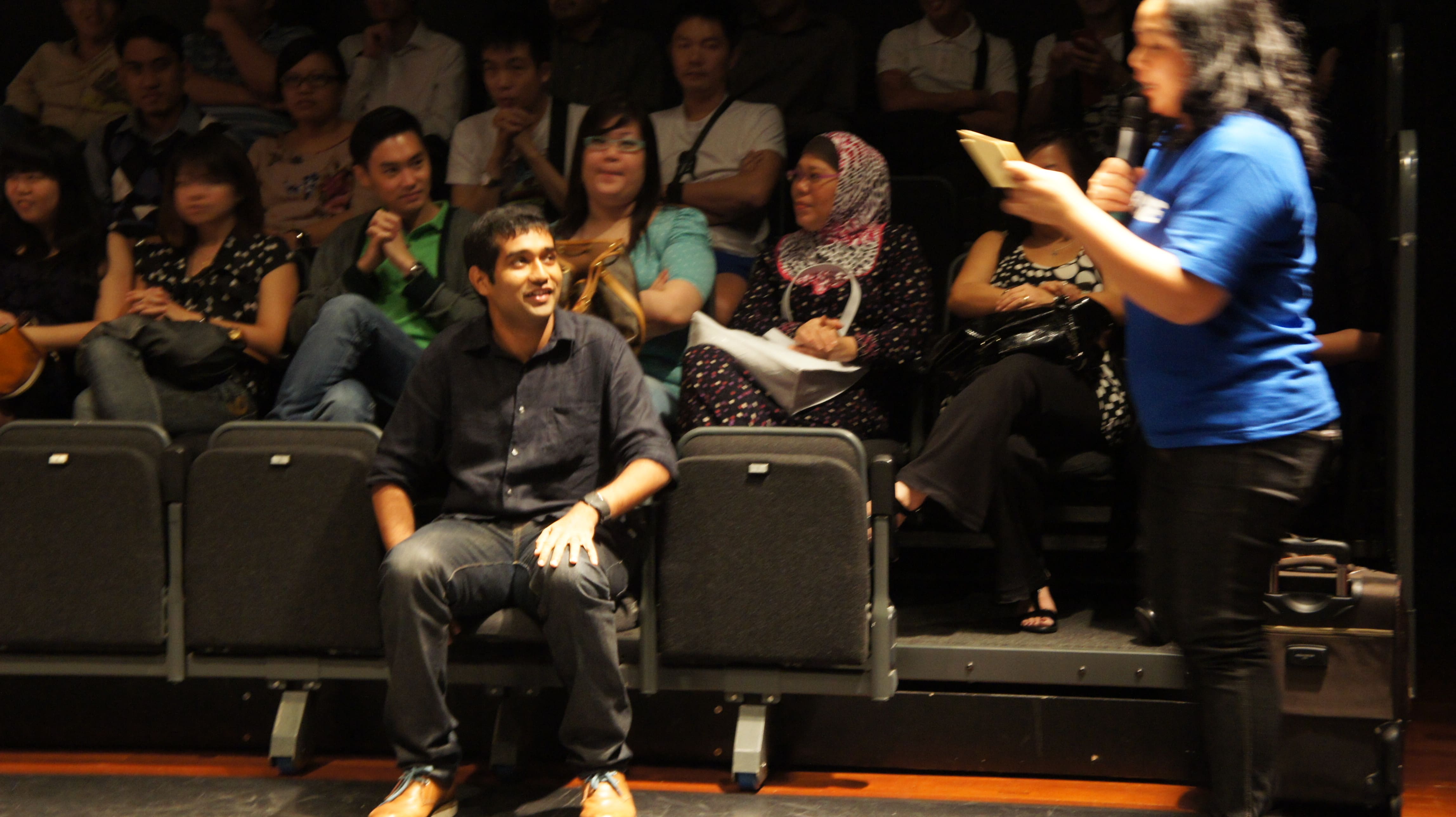 The photograph features male presenting audience member wearing black clothes, seated alone in the front row, looking at a female presenting person in a blue shirt holding a mic, reading off a piece of paper. Many more audience members are seated in the rows behind.