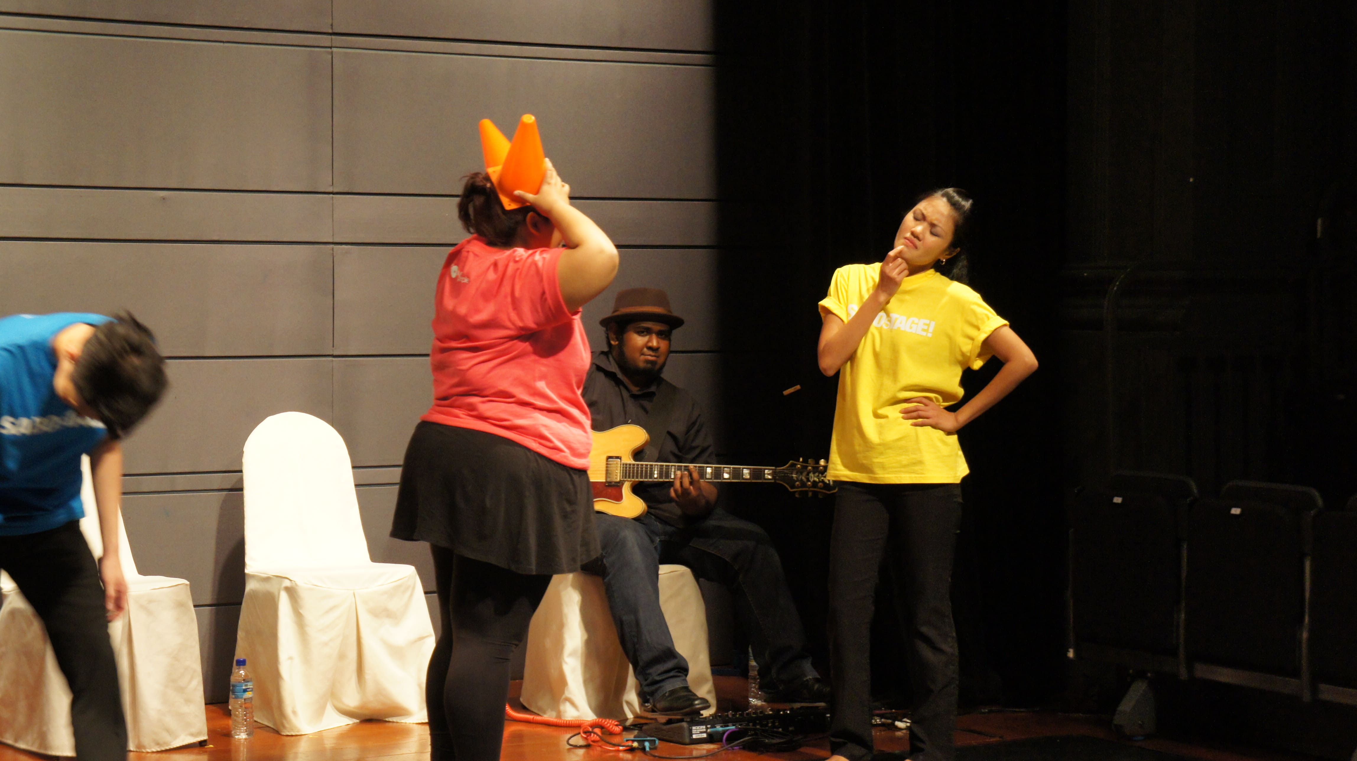 The photograph features a female presenting person wearing a pink shirt holding two orange cones on her head, while another female presenting person in yellow to the right is looking at her quizzically, with her right index finger on her chin and her left hand on her hip.