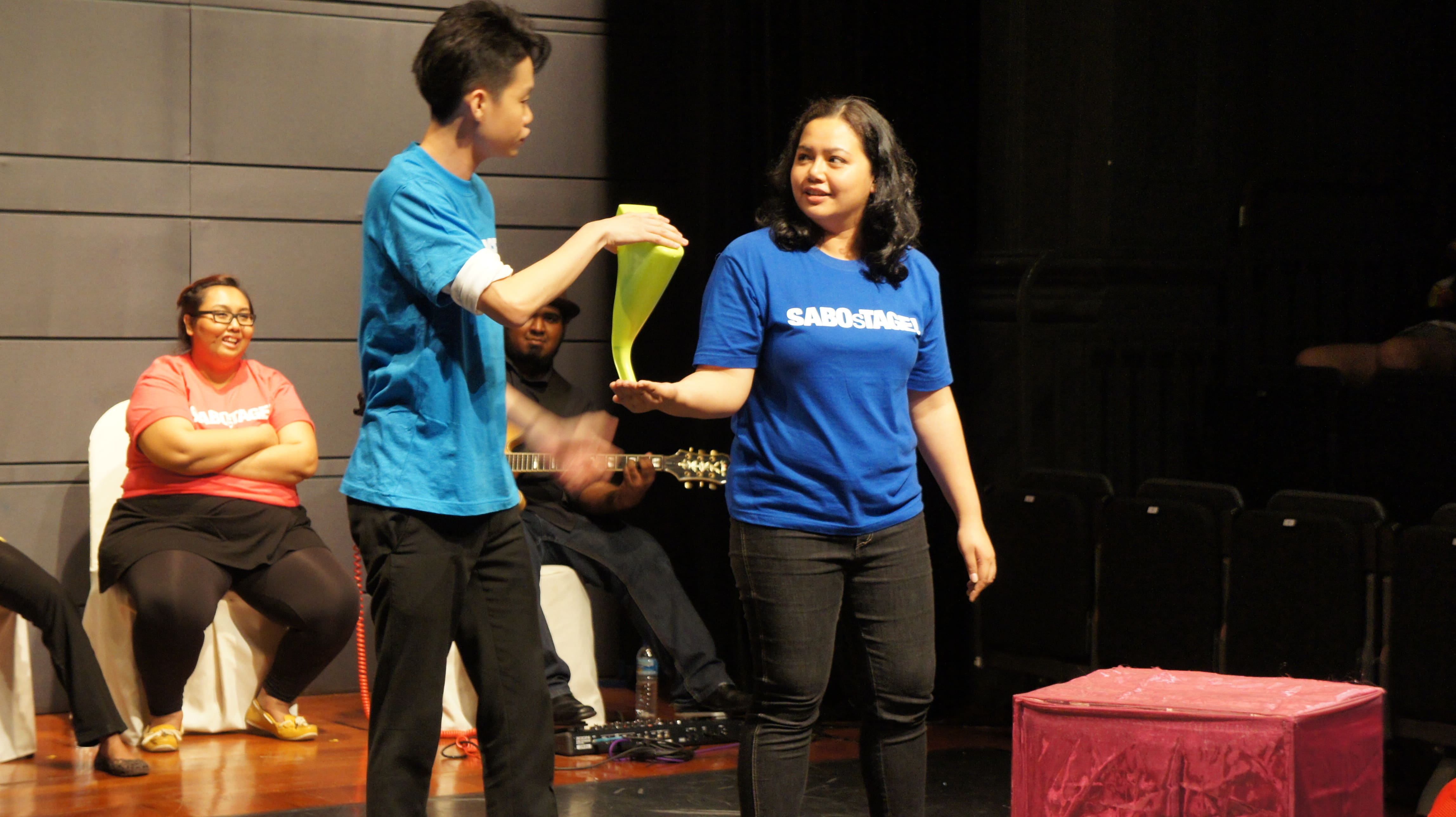 The photograph features two persons in blue shirts, holding a green object together, with the male presenting person on the left placing his right hand on top of the object, while the female presenting person on the right has her right hand supporting the bottom of the object.