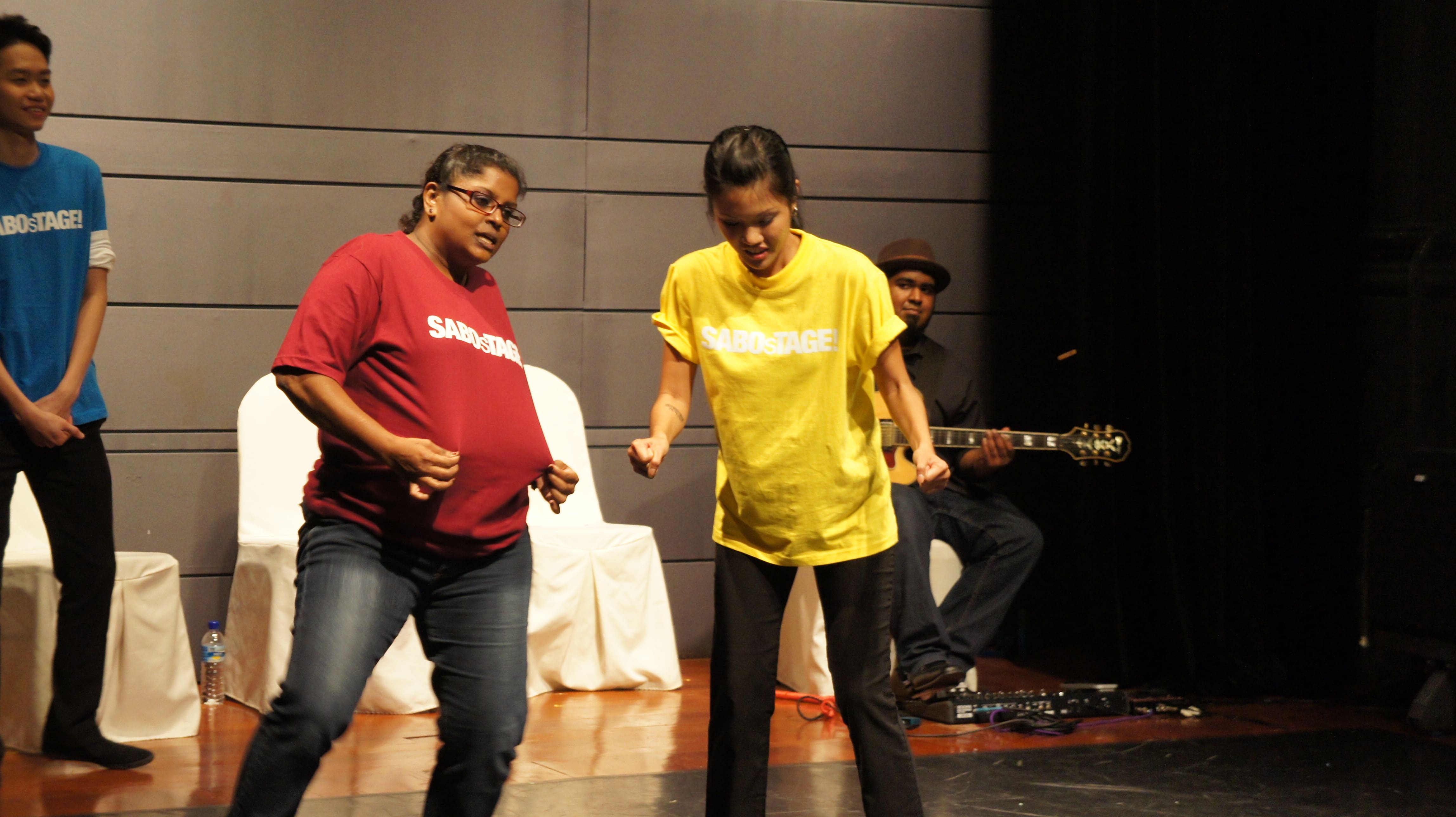 The photograph features a two female presenting persons. The one on the left is in a red shirt, and both her hands are pulling the fabric of her shirt forward, while the one on the right is in a yellow shirt, and her hands are balled into fists by her side.