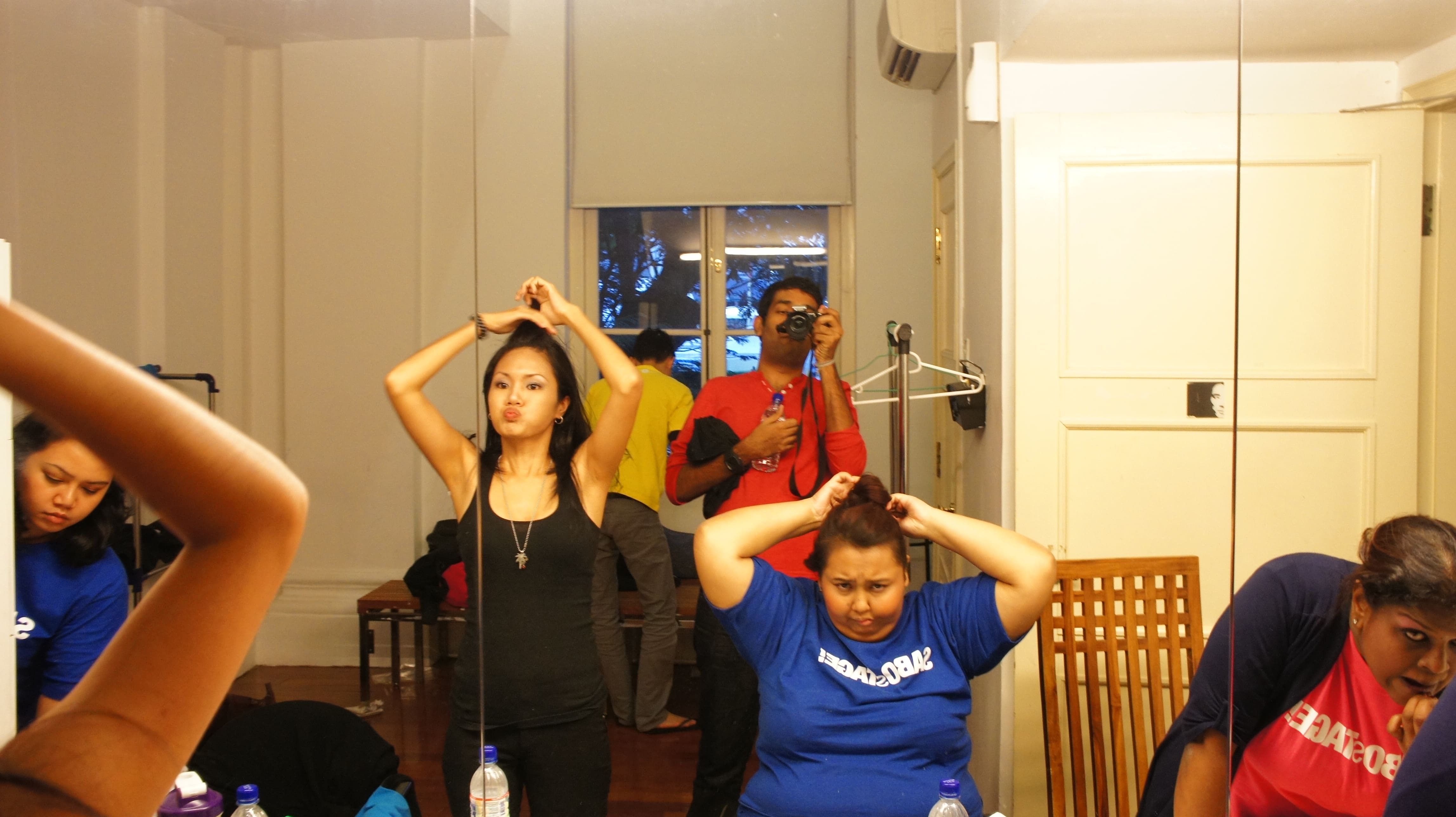 The photograph features the reflection on a dressing room mirror. Two female presenting persons can be seen in the foreground, tying up their hair, while a male presenting person holding a camera is standing behind them.