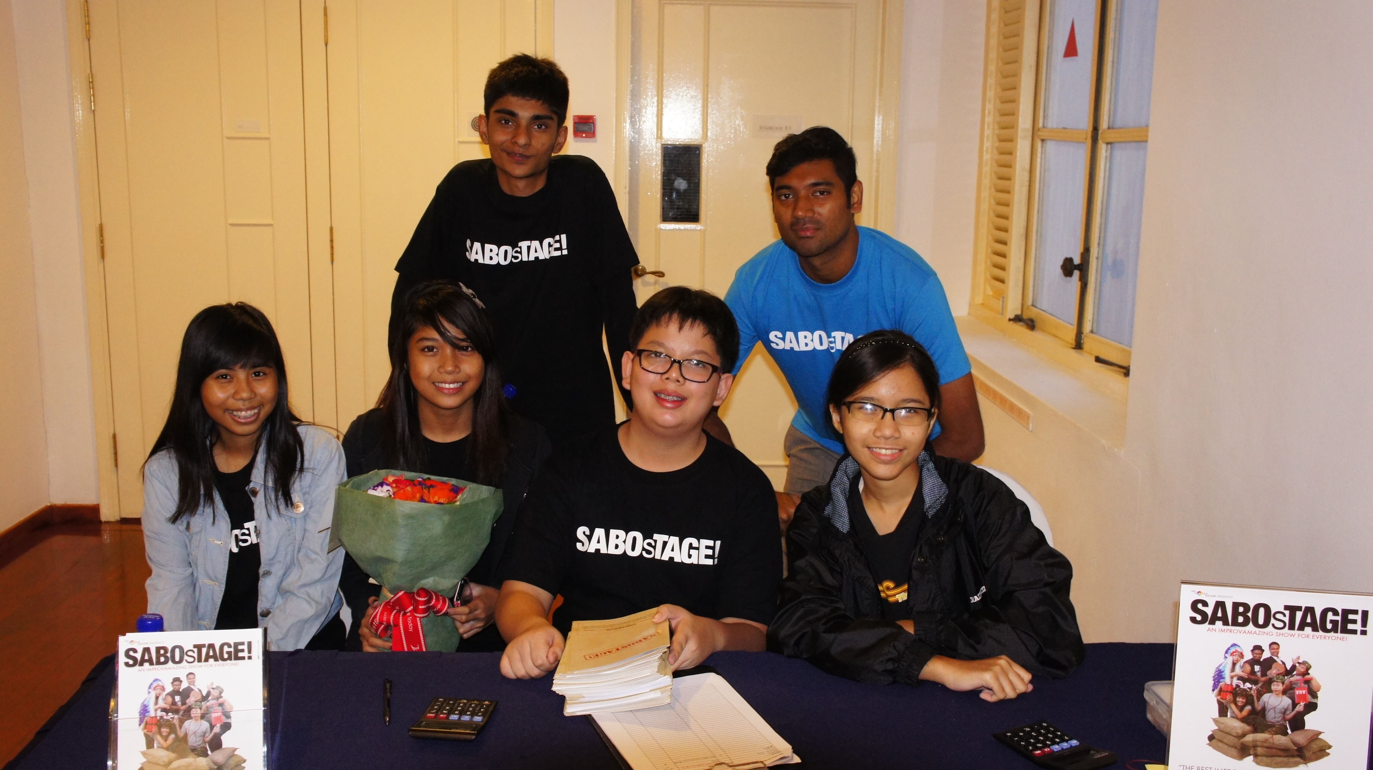 The photograph features four persons sitting behind a black table, with two more persons standing behind. Two small displays featuring the poster for "SABOsTAGE"  can be seen on the table.