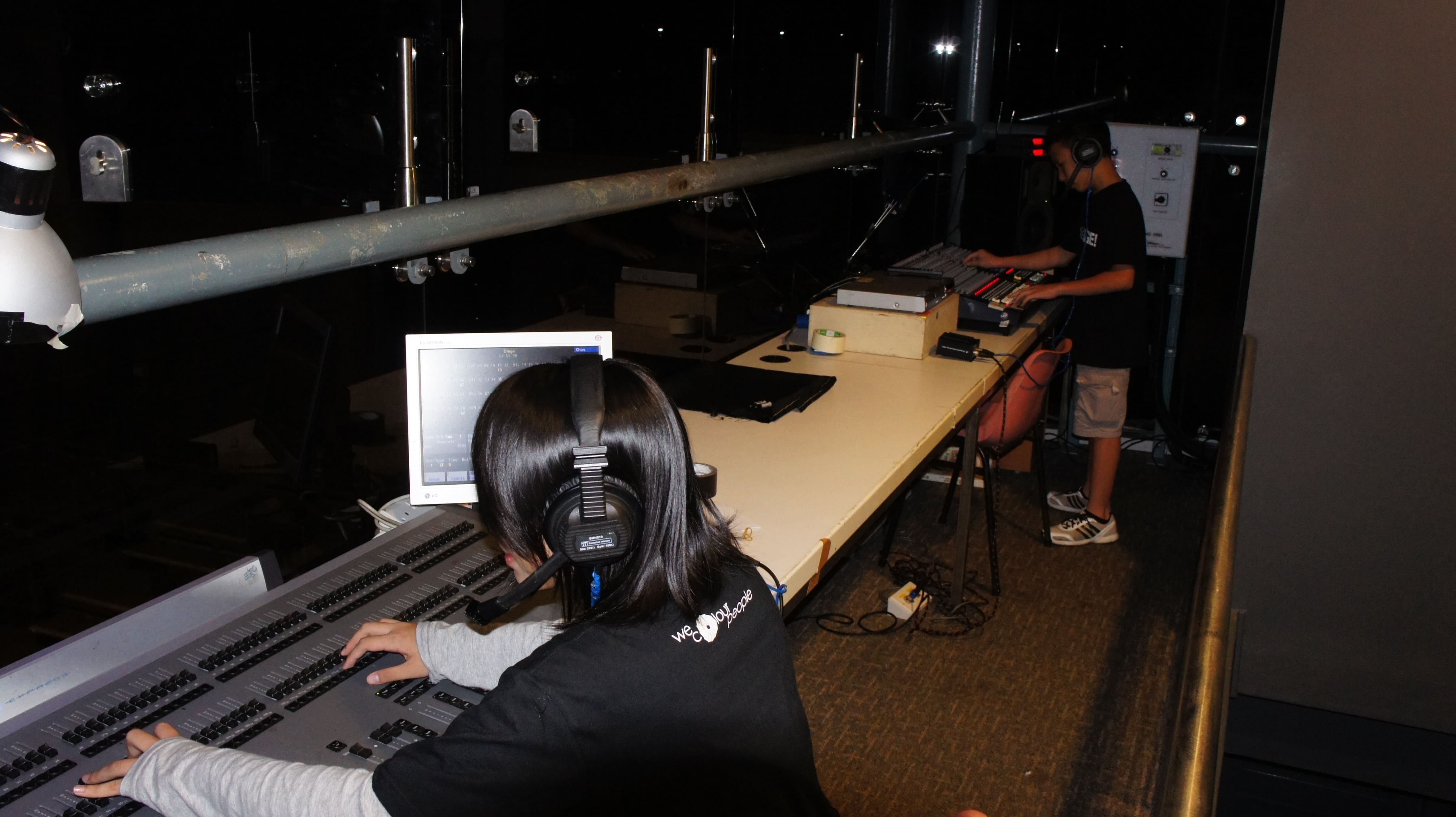 The photograph features a female presenting person on the left working on a lighting panel, while a male-presenting person is on the right, operating a sound board.