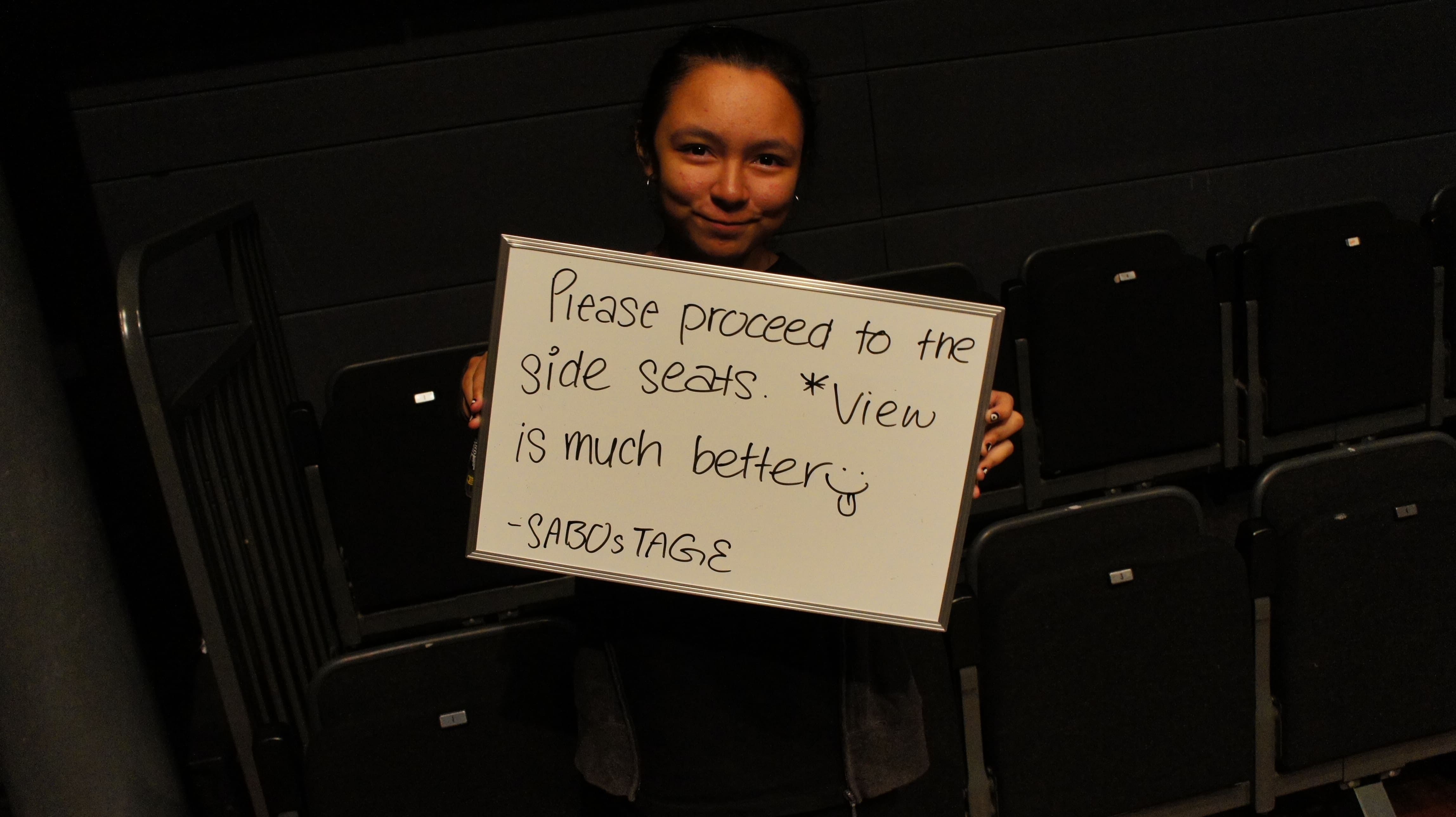 The photograph features a female presenting person in front of audience seats, holding a whiteboard that says "Please proceed to the side seats. *View is much better. - Sabostage".