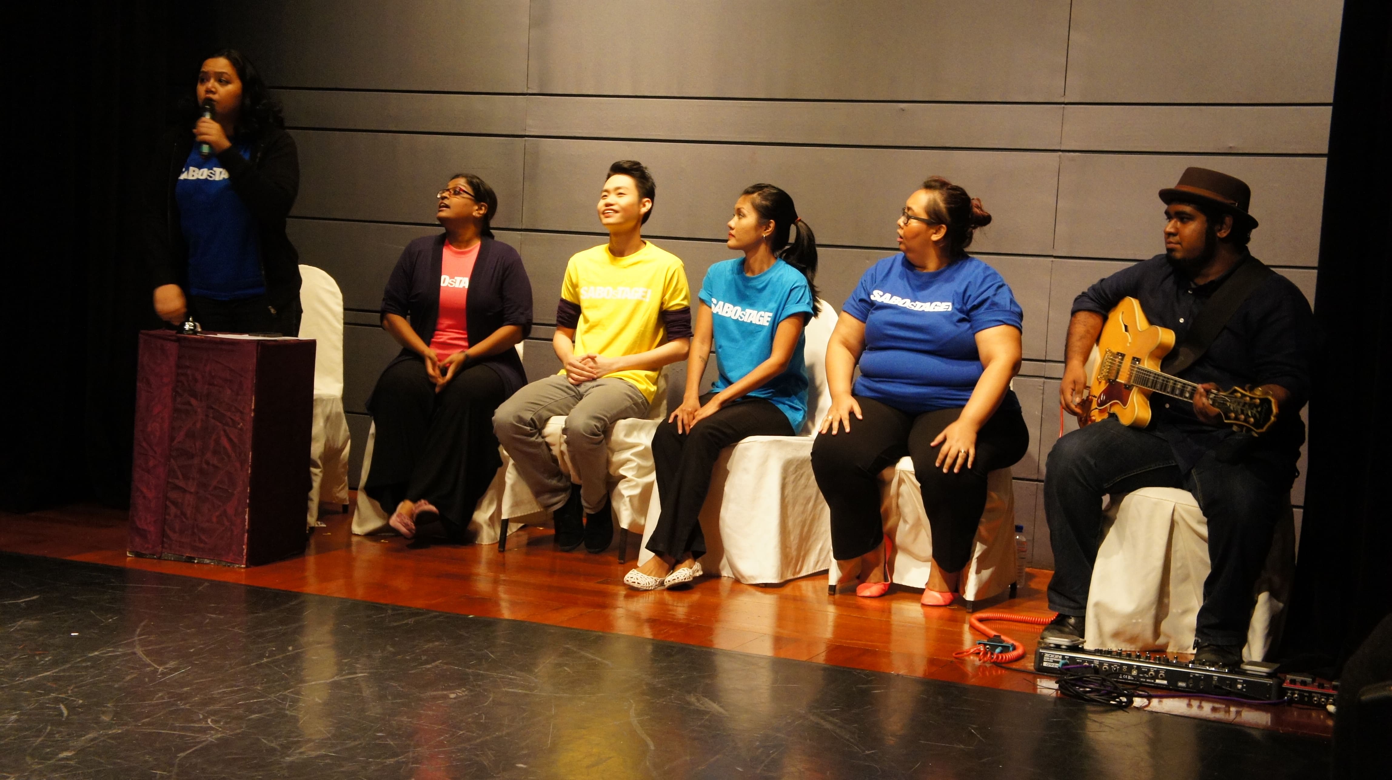 The photograph features a female presenting person wearing a blue SABOsTAGE shirt on the far left standing and speaking at a podium. To her right, four people in SABOsTAGE shirts of varying colours are seated on white chairs, while a male presenting person in black and wearing a hat is seated on the far right holding a guitar.