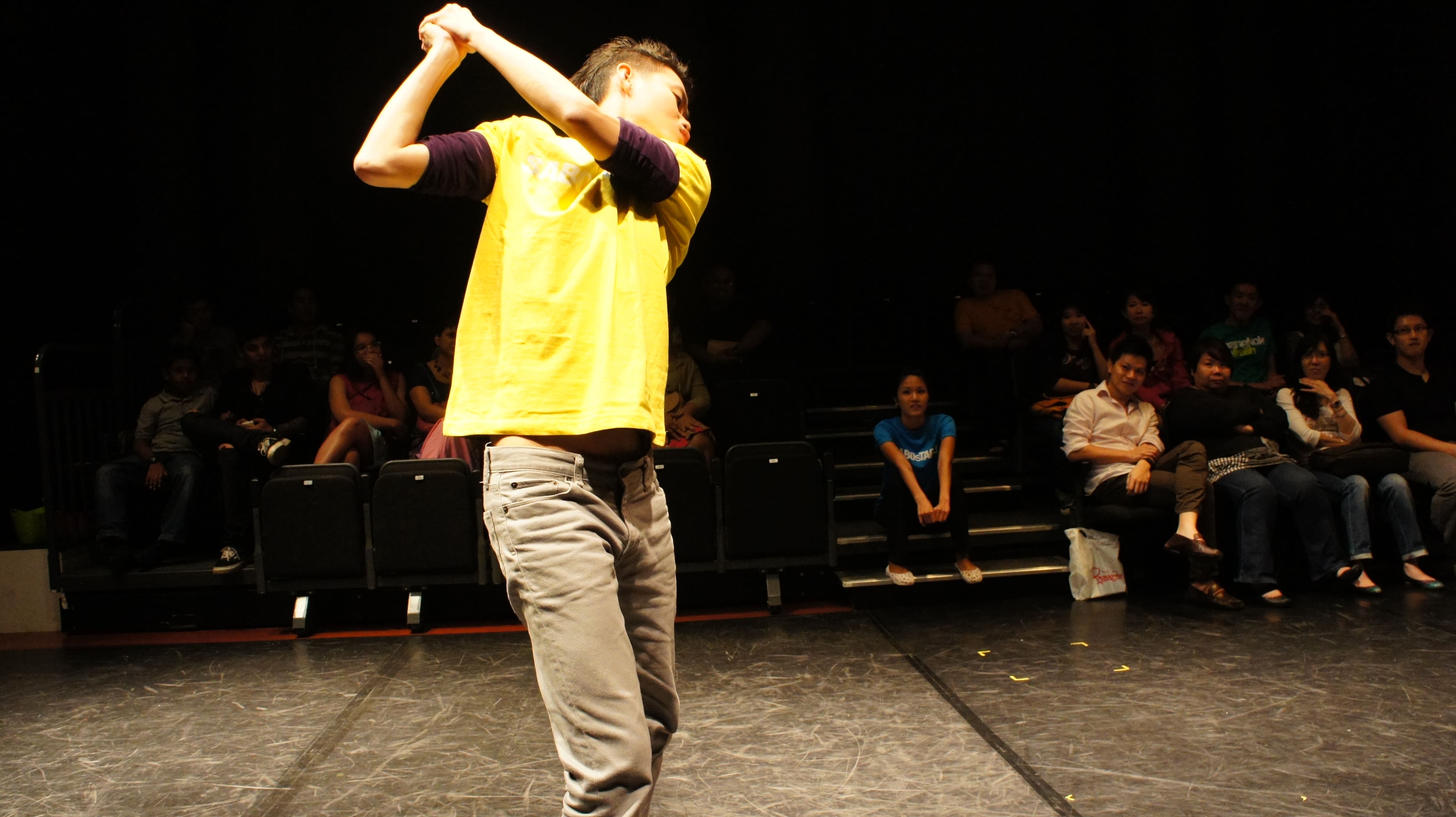 The photograph features a male presenting person in a yellow SABOsTAGE shirt, miming a golf swing towards the audience, while a female presenting person in a blue SABOsTAGE shirt sits on the steps next to audience seating.