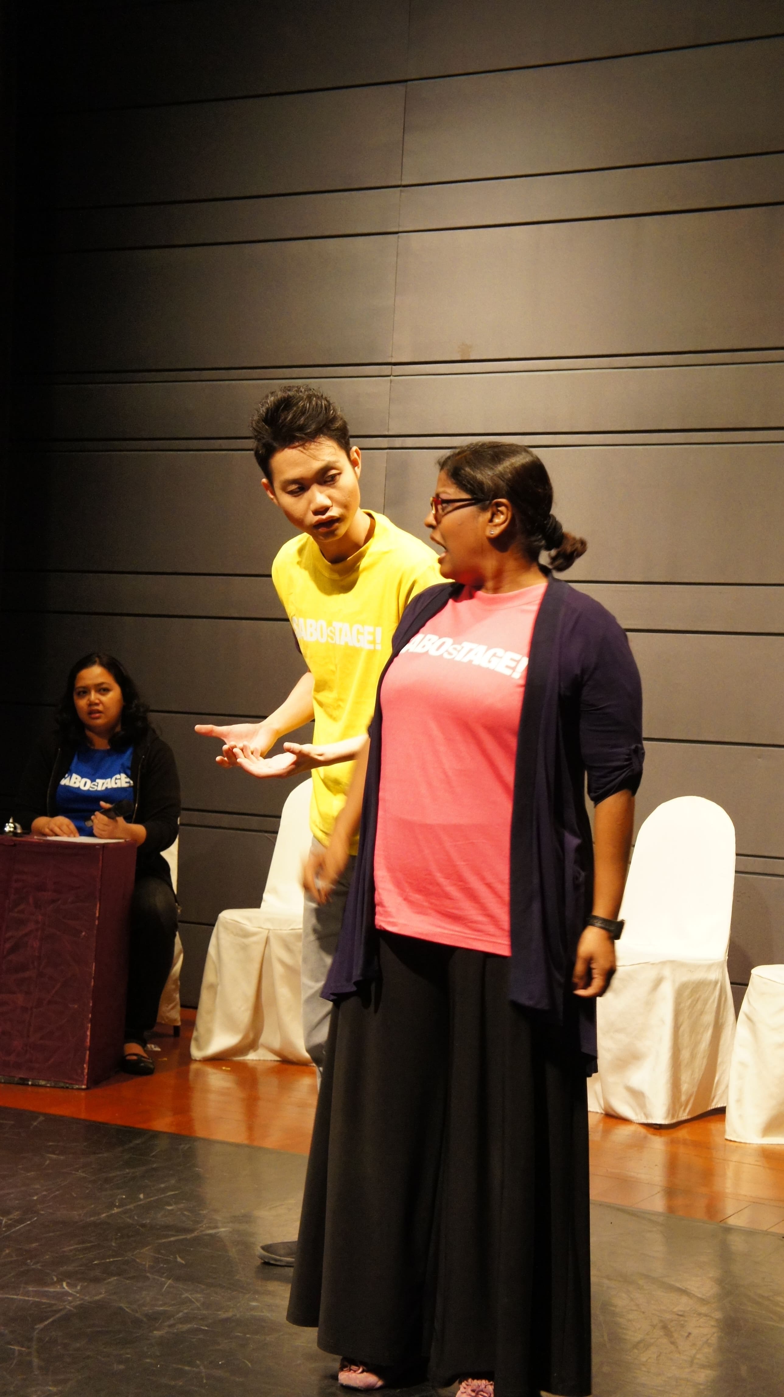 The photograph features a male presenting person in a yellow SABOsTAGE shirt with his right hand slightly outstretched, speaking to a female-presenting person in a pink SABOsTAGE shirt to his left, who is turning to face him.