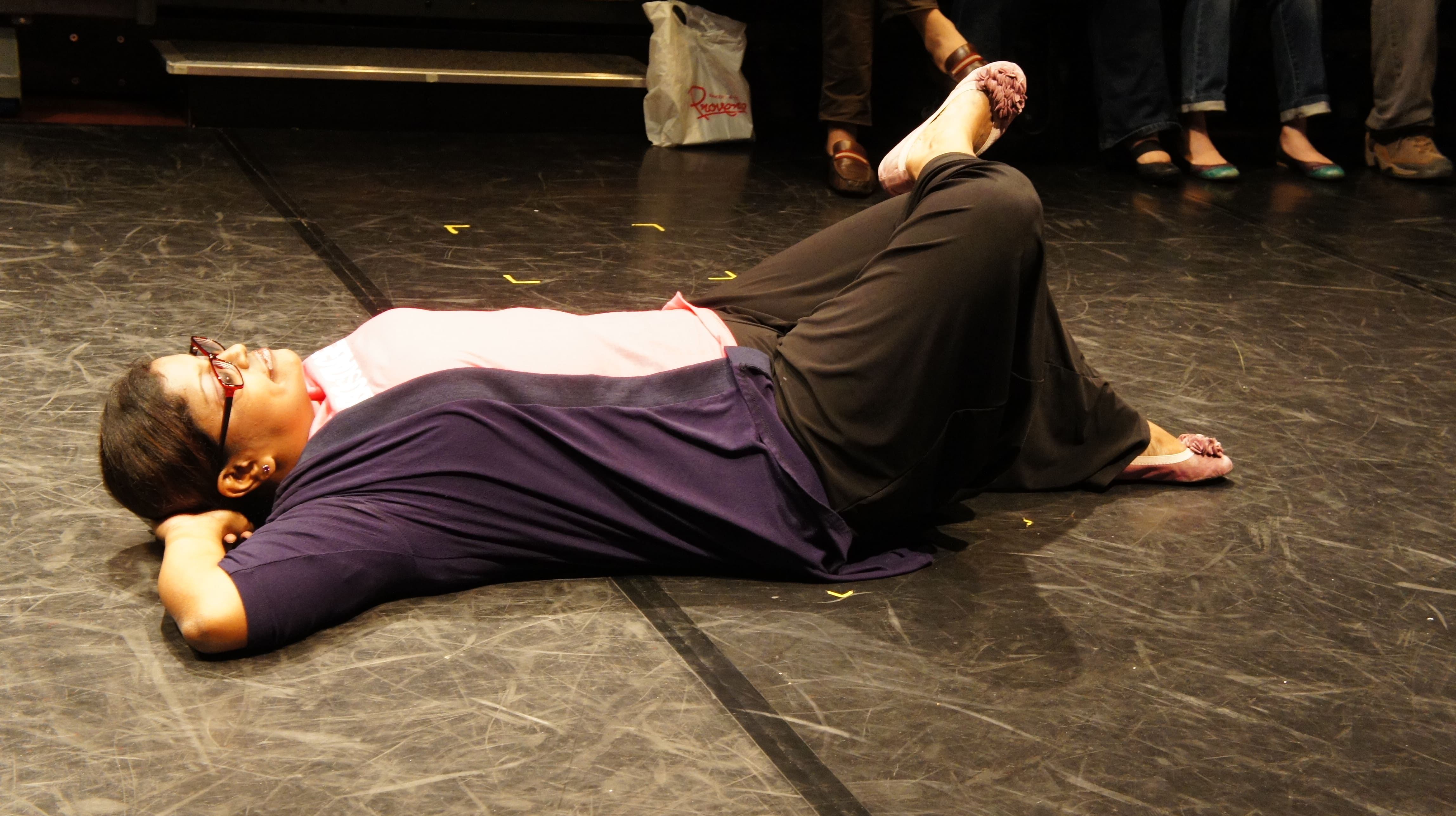 The photograph features a female presenting person in a pink shirt and a blue jacket, lying on the ground with her hands under her head and her right leg propped up on her left knee.