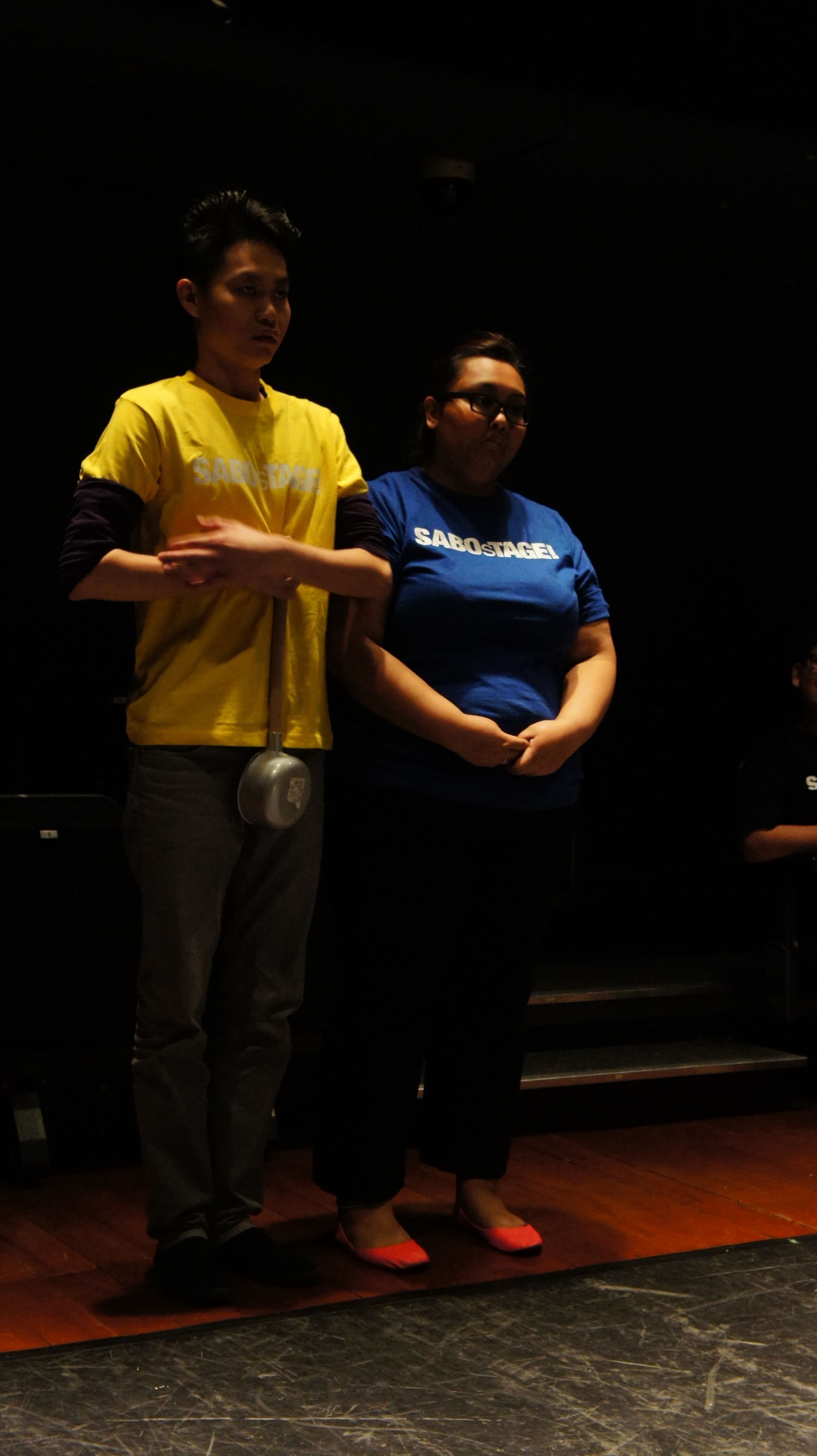 The photograph features a male presenting person in a yellow SABOsTAGE shirt with his arms across his upper abdomen, holding a large ladle in his right hand, pointed downwards. A female person in a blue SABOsTAGE shirt is to his left, with her hands clasped in front of her lower abdomen.