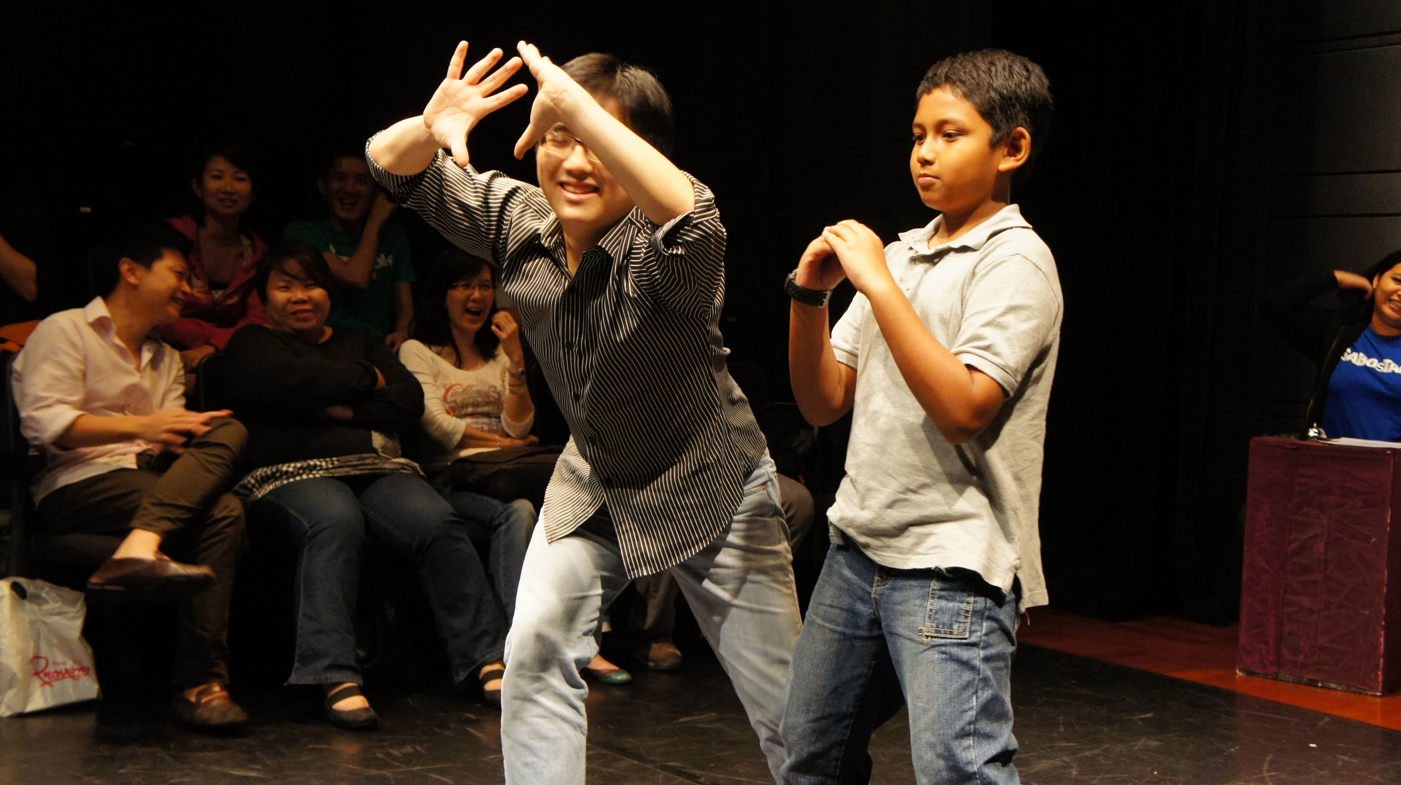 The photograph features two male presenting persons, one adult and one child. The adult is on the left, leaning forward with his arms situated in front of his head, palms open and facing away from him, while the child is on the right, his hands clasped in front of his chest. The audience in the background can be seen smiling and laughing.