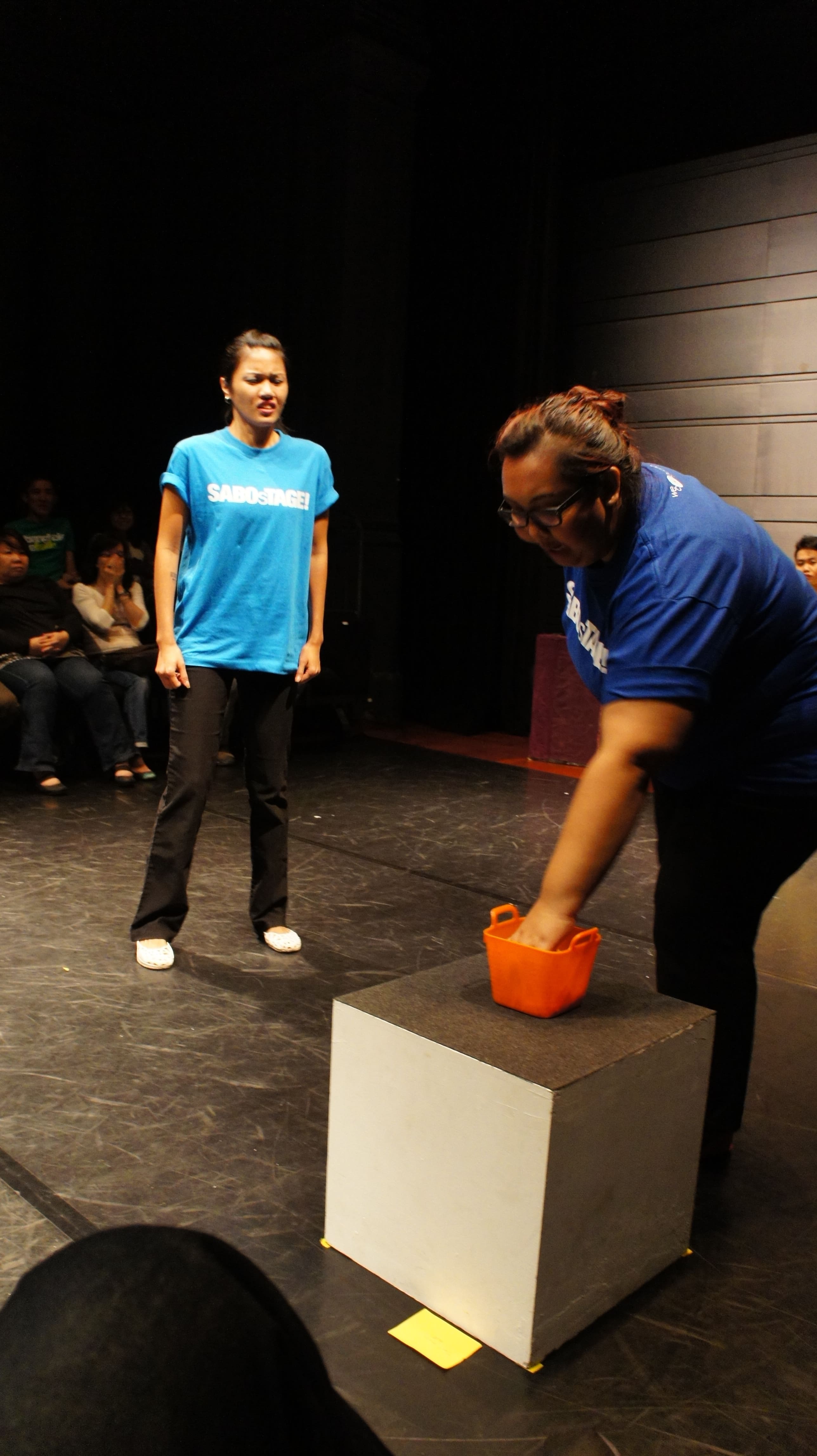 The photograph features a female presenting person wearing a dark blue SABOsTAGE shirt in the foreground, reaching her left hand down into an orange container situated on a cube, while another female presenting person in a light blue SABOsTAGE shirt is standing in the background, arms by her side, frowning at her.