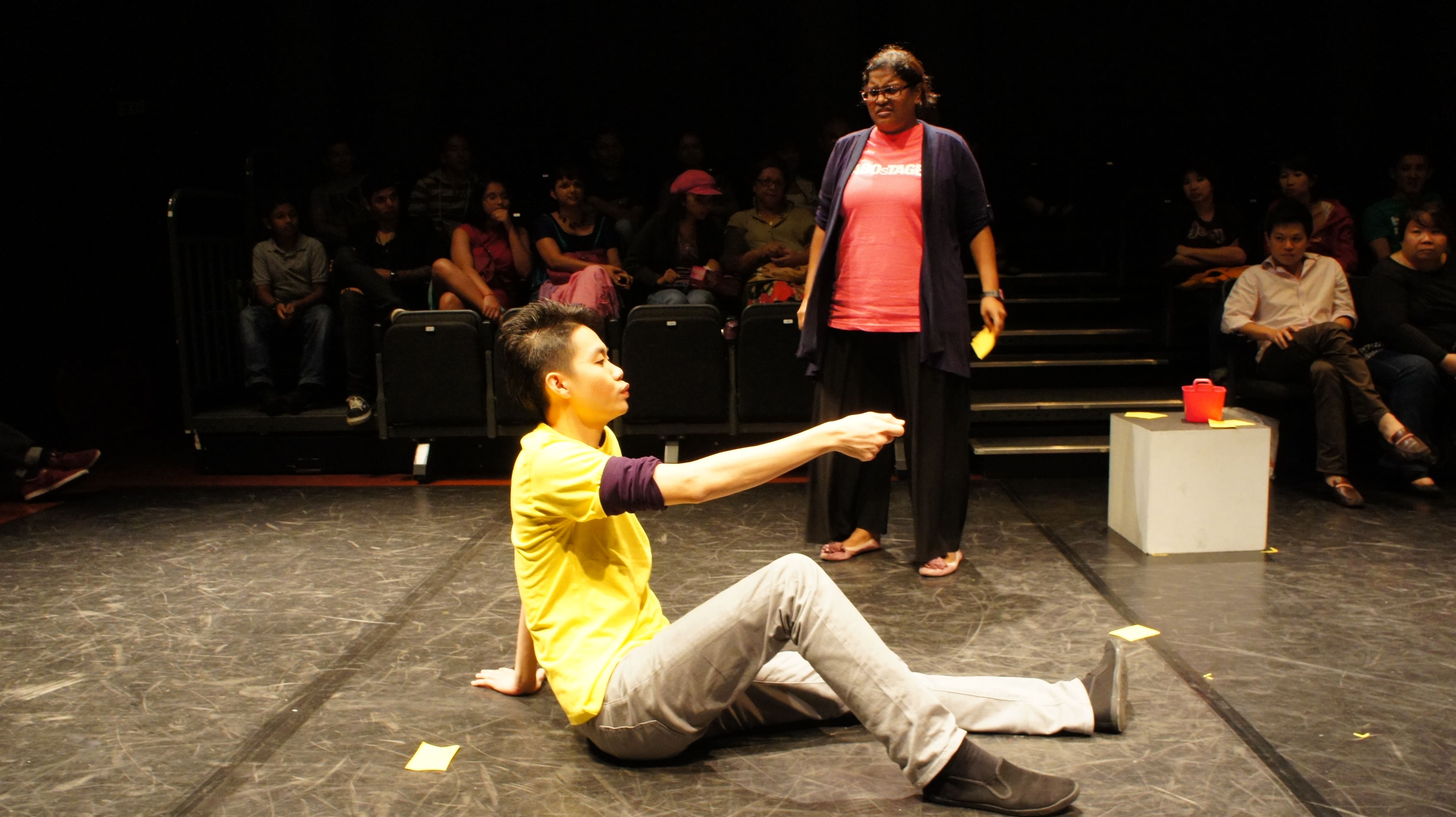 The photograph features a male presenting person in a yellow SABOsTAGE shirt in the foreground, seated with one leg bent and one leg straightened, pointing his right arm towards the right of the photo, while a female presenting person in a pink SABOsTAGE shirt stand in the background, arms to her side, frowning at him.