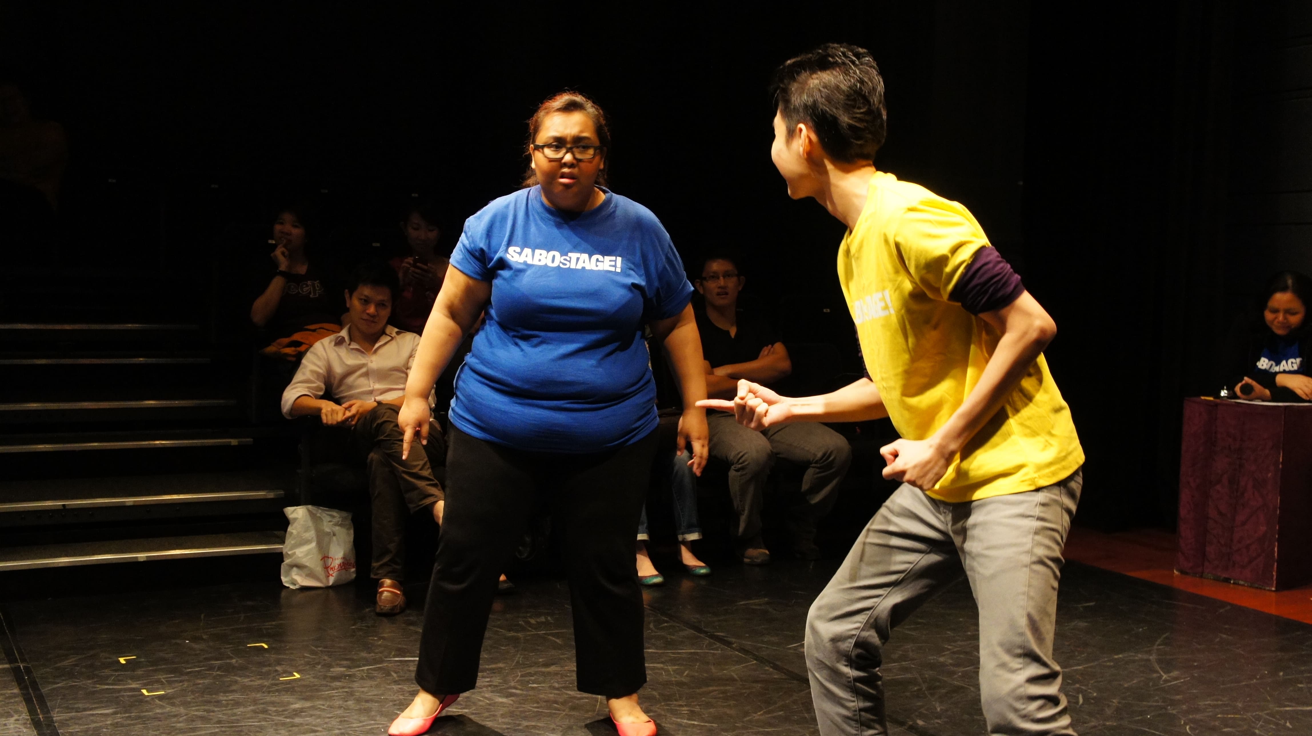 The photograph features a female presenting person in a dark blue SABOsTAGE shirt, standing with her arms to her side, leaning slightly towards and frowning at a male presenting person wearing a yellow SABOsTAGE shirt, who is leaning towards the left of the photo, with his right arm pointed towards the left as well.