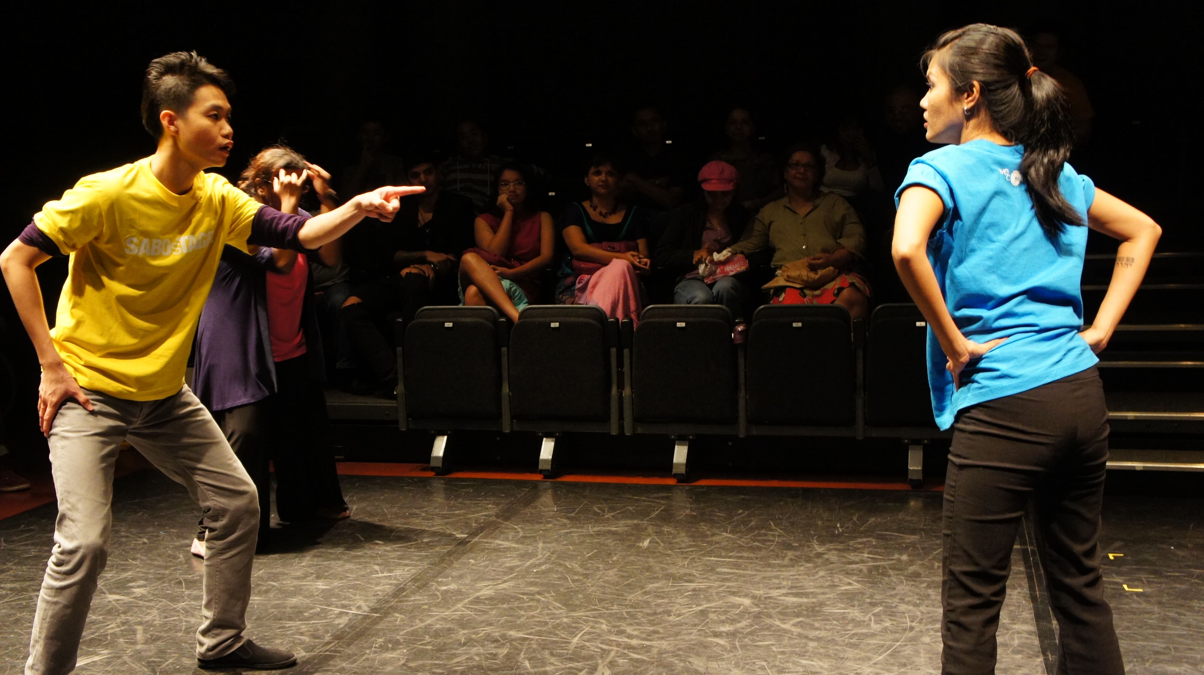 The photograph features a male presenting person in a yellow SABOsTAGE shirt on the left, pointing towards a female presenting person in a light blue shirt on the right, who has her hands on her hips.