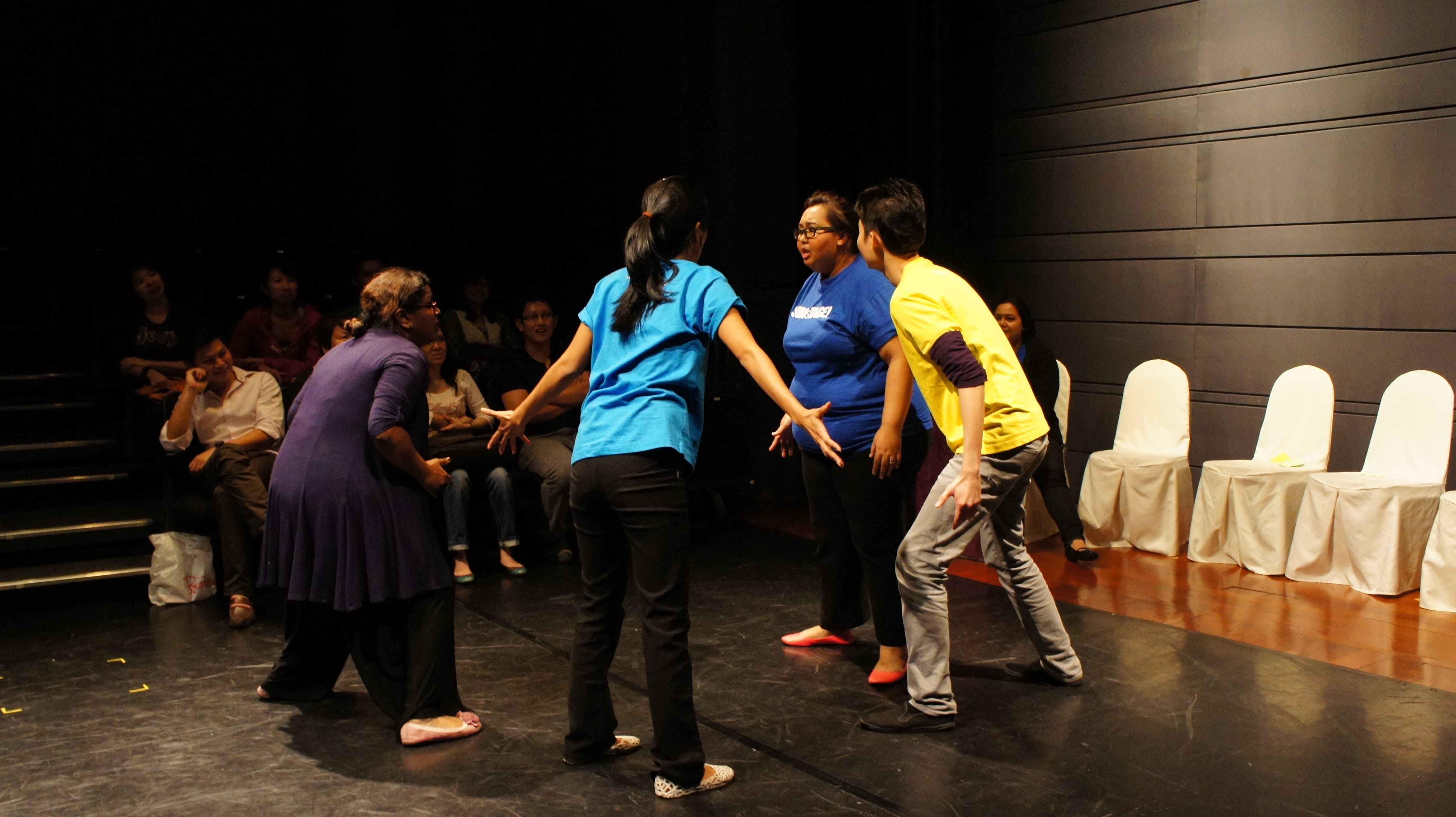 The photograph features four persons in shirts of various colours, standing in a circle facing each other. Three of them have are leaning slightly into the circle, with their arms by their side, while the one in the centre and closest to the camera has her hands stretched diagonally downwards to her sides.