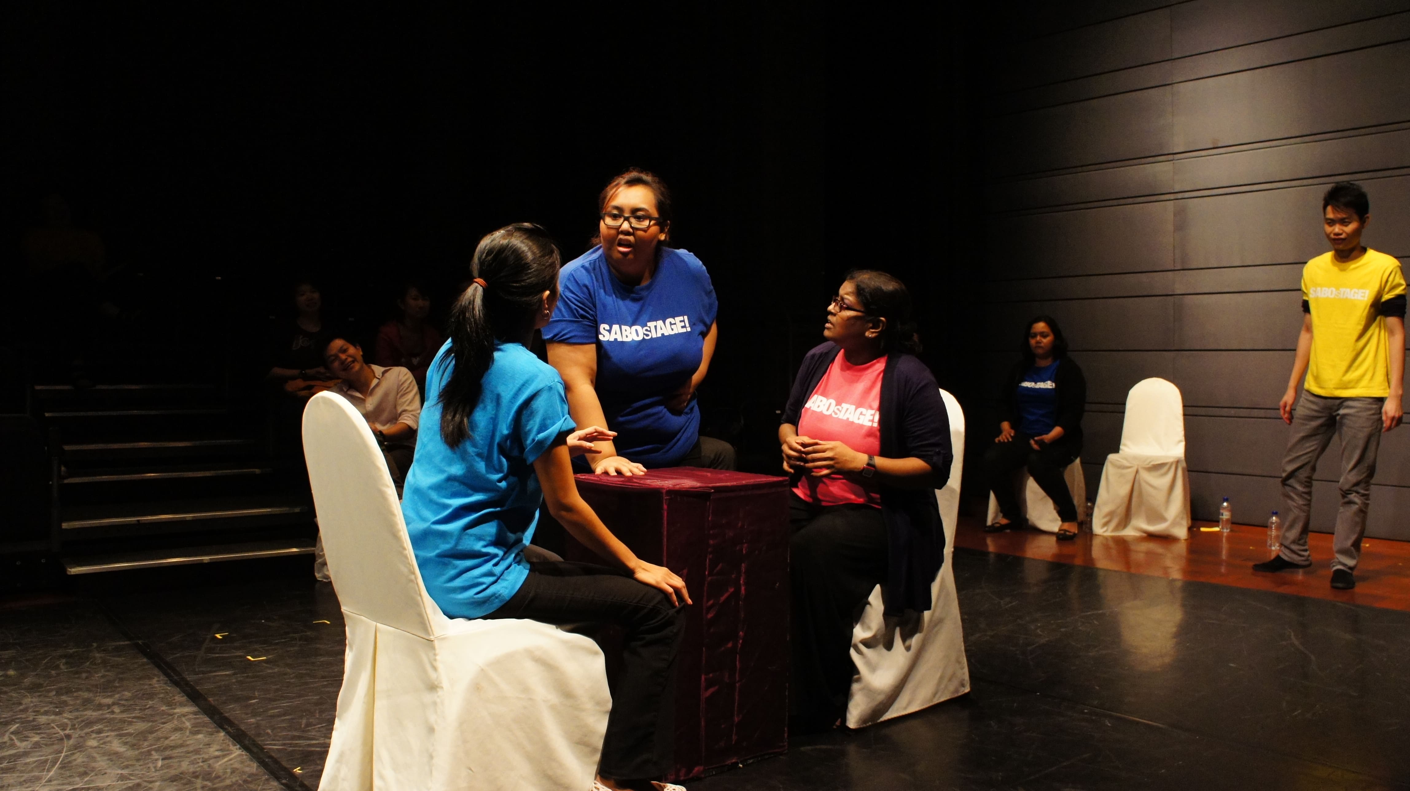 The photograph features two female presenting persons sitting at a square table, one in a light blue shirt and another in a pink SABOsTAGE shirt. A third female presenting person in a dark blue SABOsTAGE shirt is standing behind the table speaking, with her right hand placed on the tabletop.