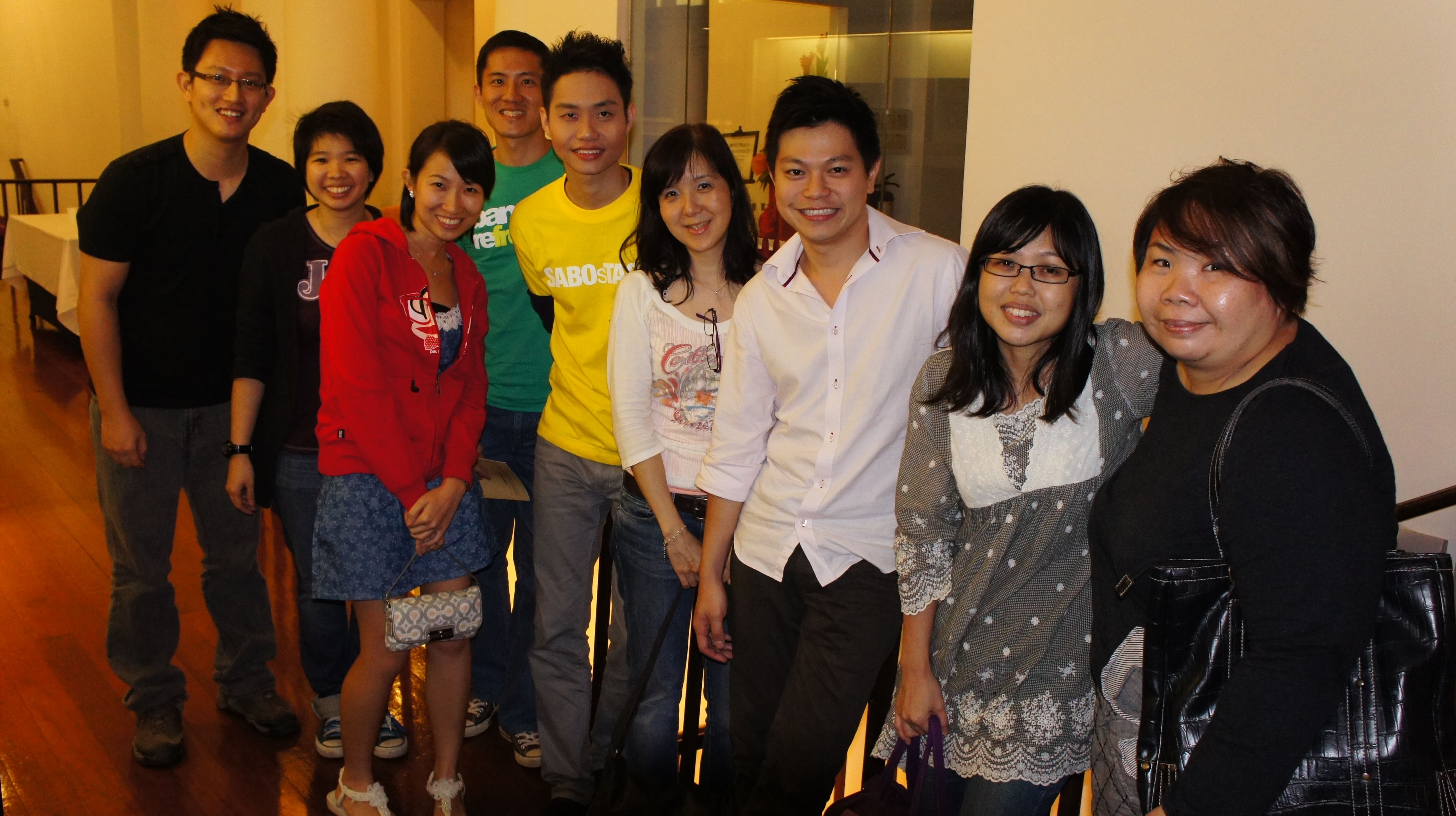 The photograph features nine persons in various attires, standing in a line along a railing, all smiling at the camera.