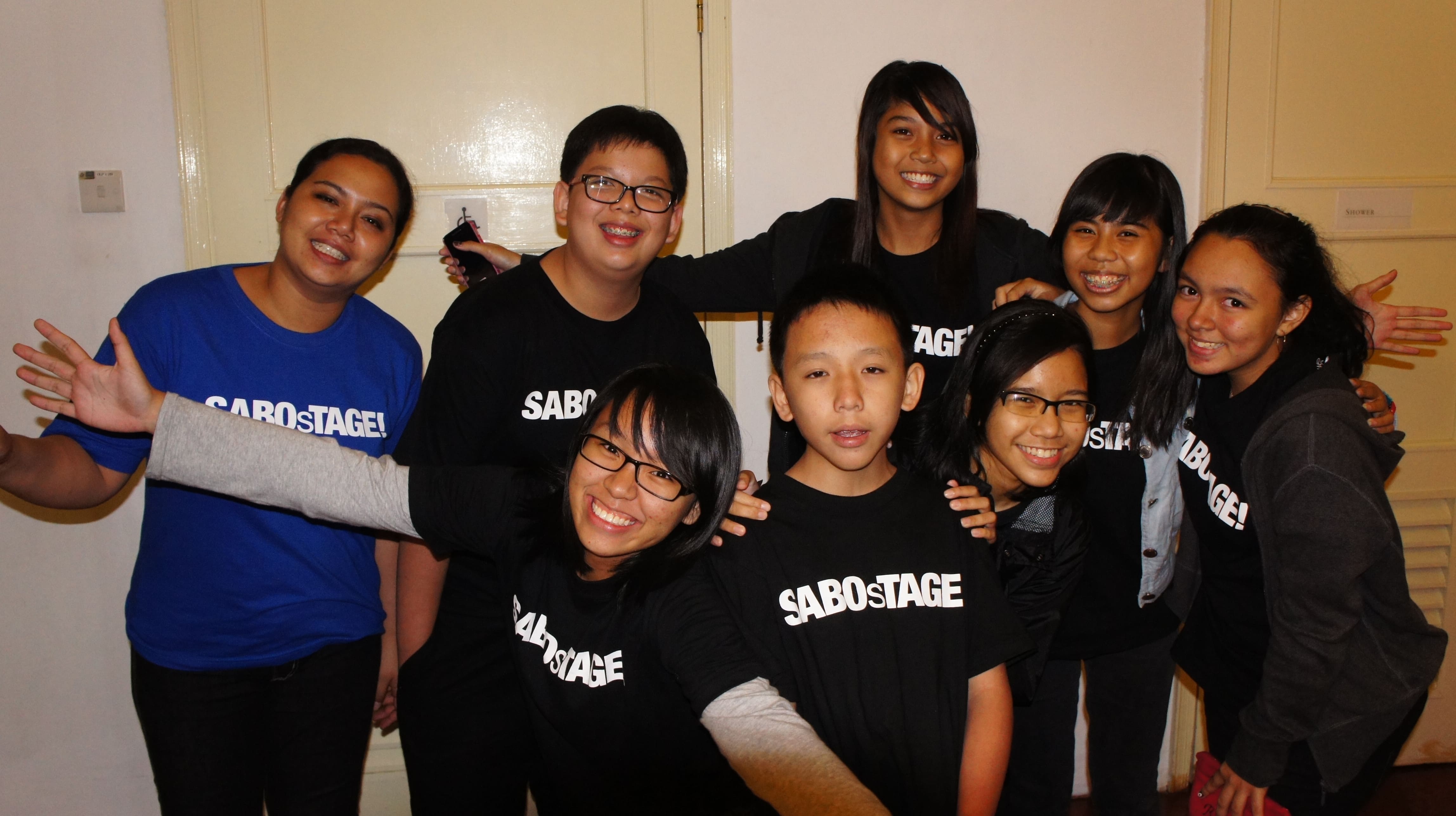 The photograph features eight persons arranged in two rows in front of two cream coloured doors, smiling at the camera. Seven of them are in black SABOsTAGE shirts, while the one on the furthest left is wearing a dark blue SABOsTAGE shirt.