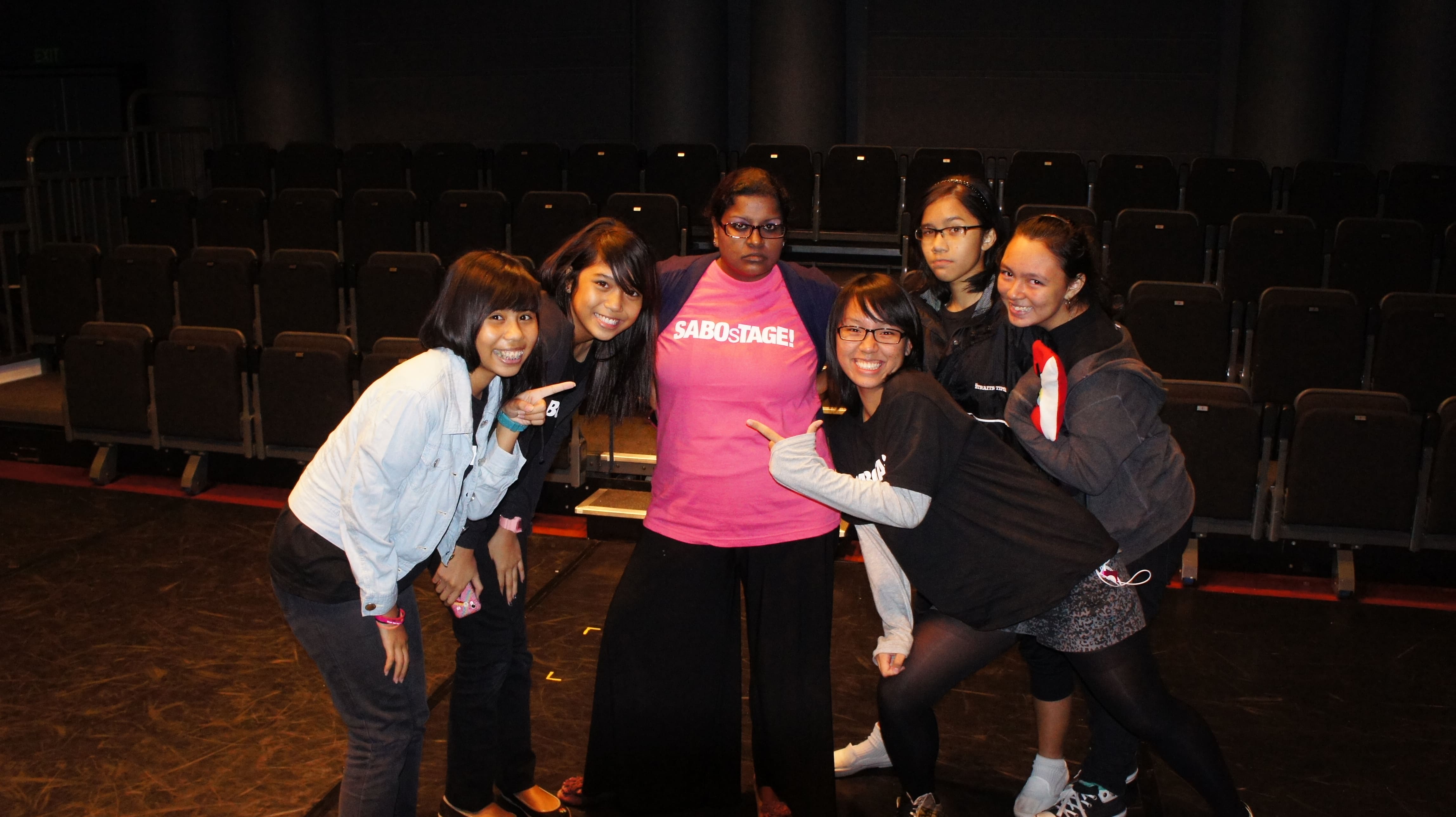 The photograph features six female presenting persons in various attires, arranged in a U shape with the opening facing the camera. The two persons furthest from the camera have serious expression on their face, while the rest are smiling into the camera.