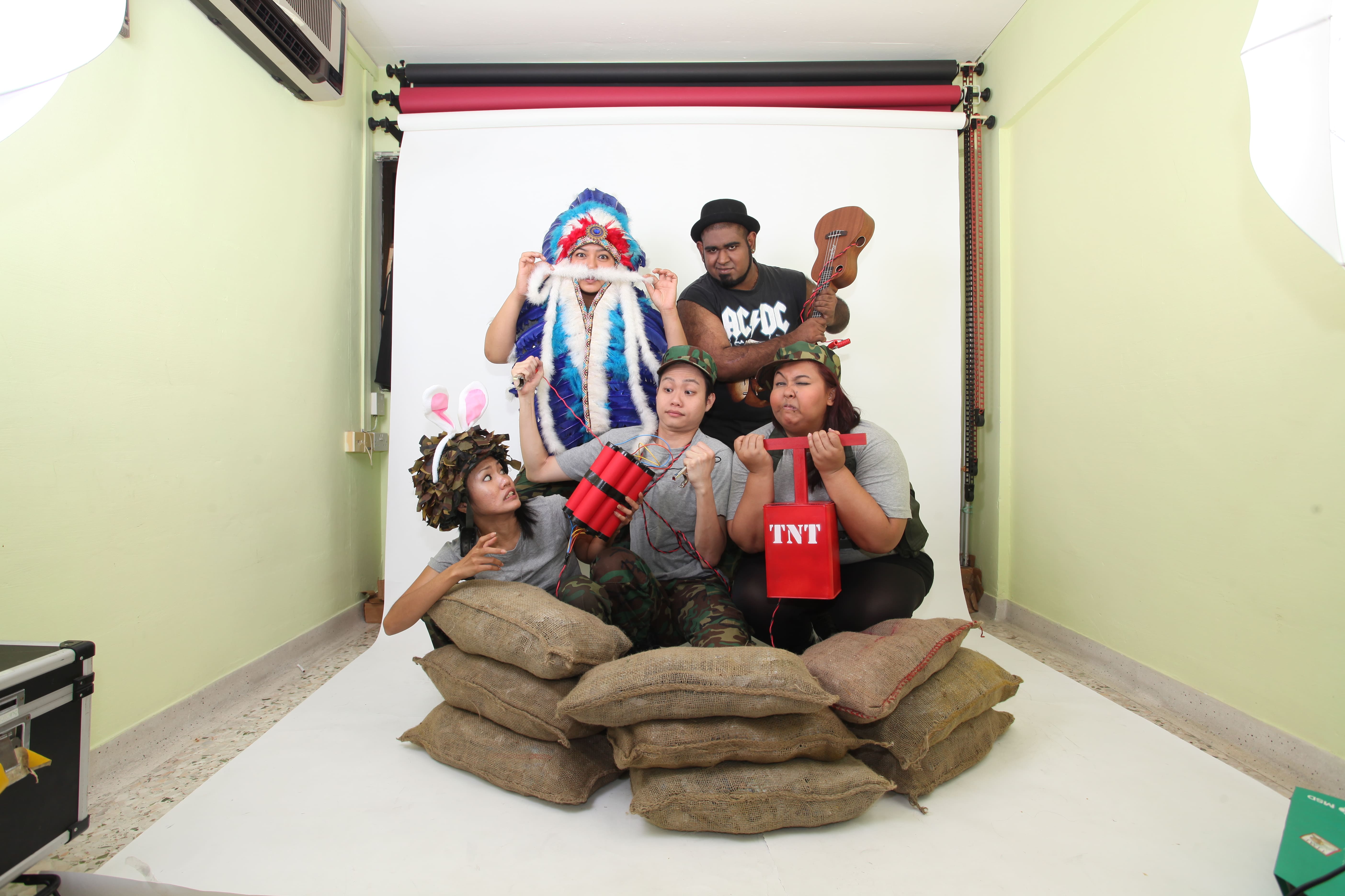 The photograph features five individuals in various comical attires against a white paper backdrop, behind a line of sandbags. They are interacting with various props, including a bundle of dynamite and a detonator, a pair of jumper cables, a fake white moustache, and a ukelele.