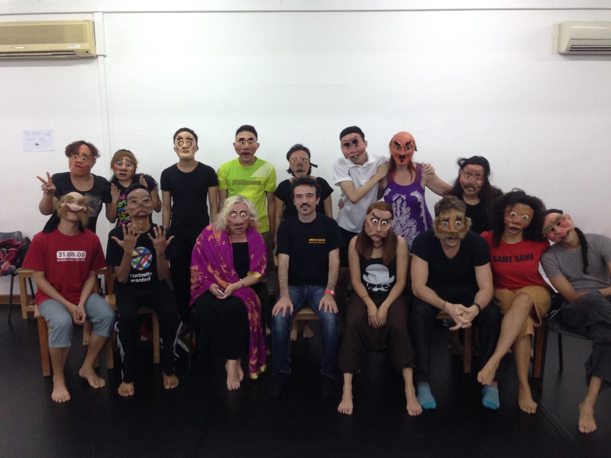 The photograph features sixteen persons arranged in two rows with eight individuals per row. The persons are dressed in various attires, and all of them are wearing masks of varying designs except the individual in the middle of the first row, who is unmasked.