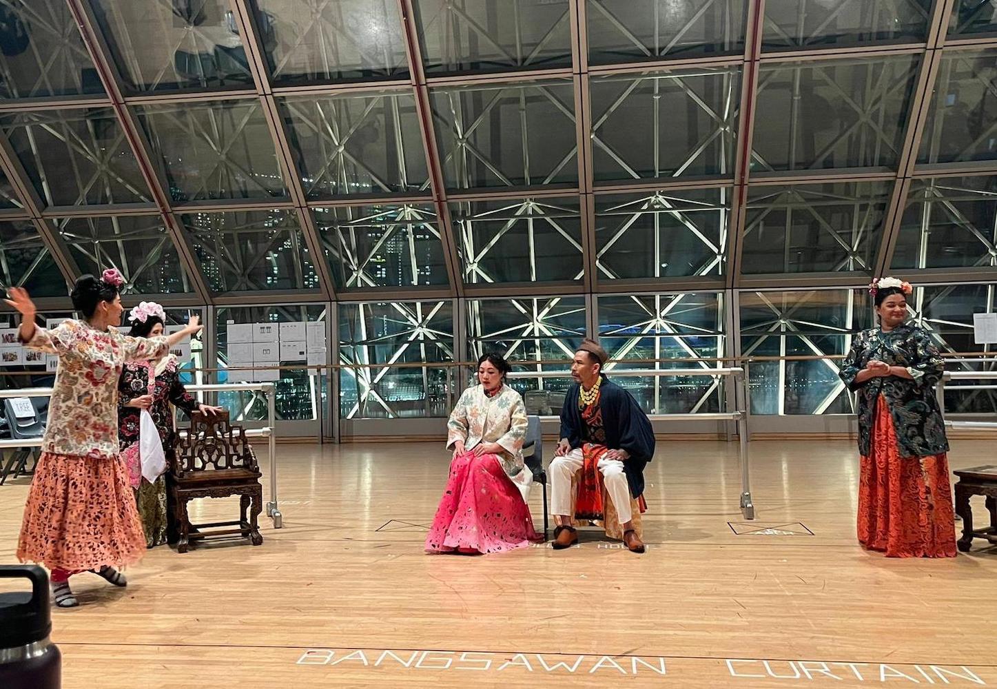 A female-presenting person gesturing widely at two persons sitting on chairs watching her.