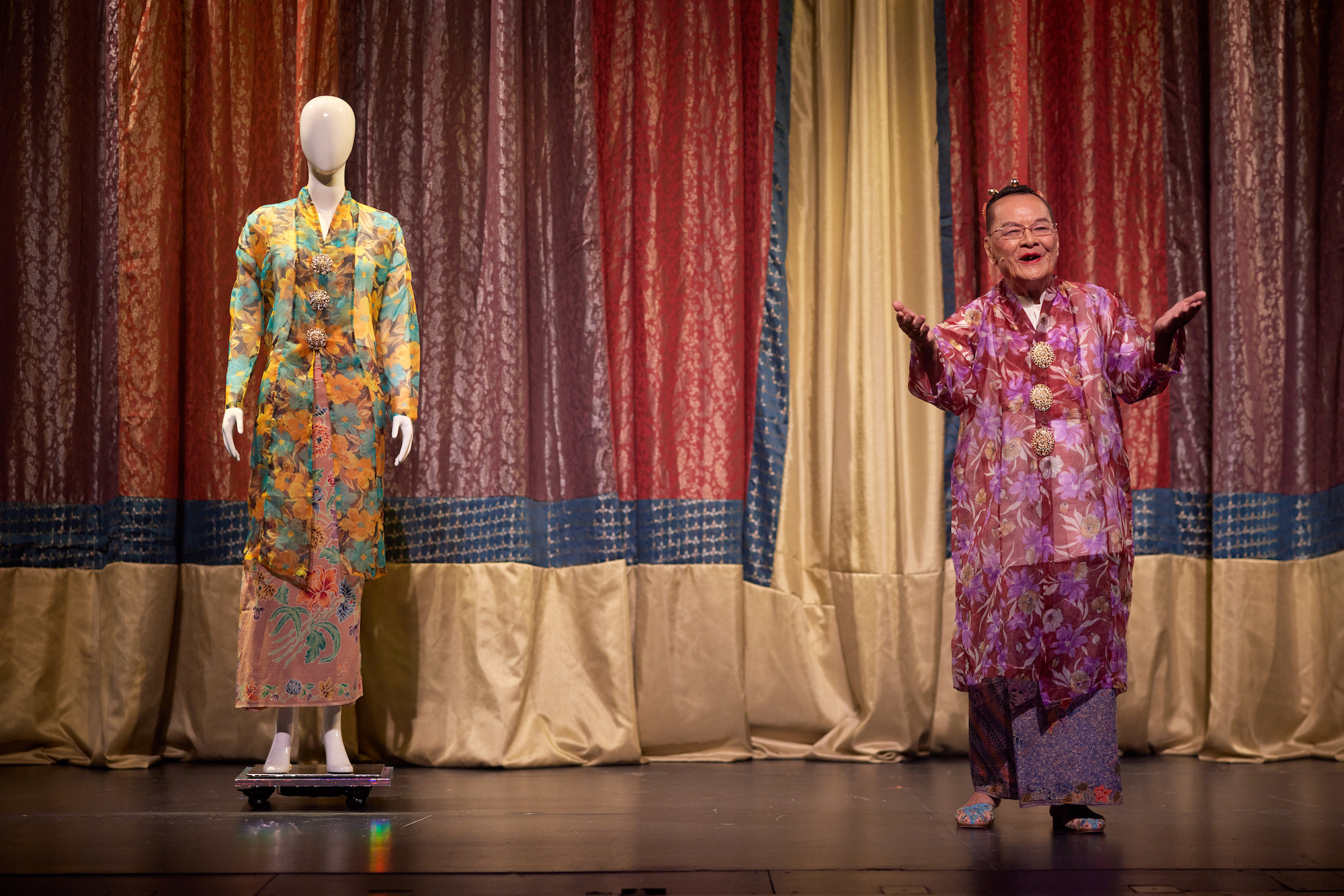 A person in elaborate pink costume, hands out and smiling. To their right is a mannequin wearing a colourful outfit.