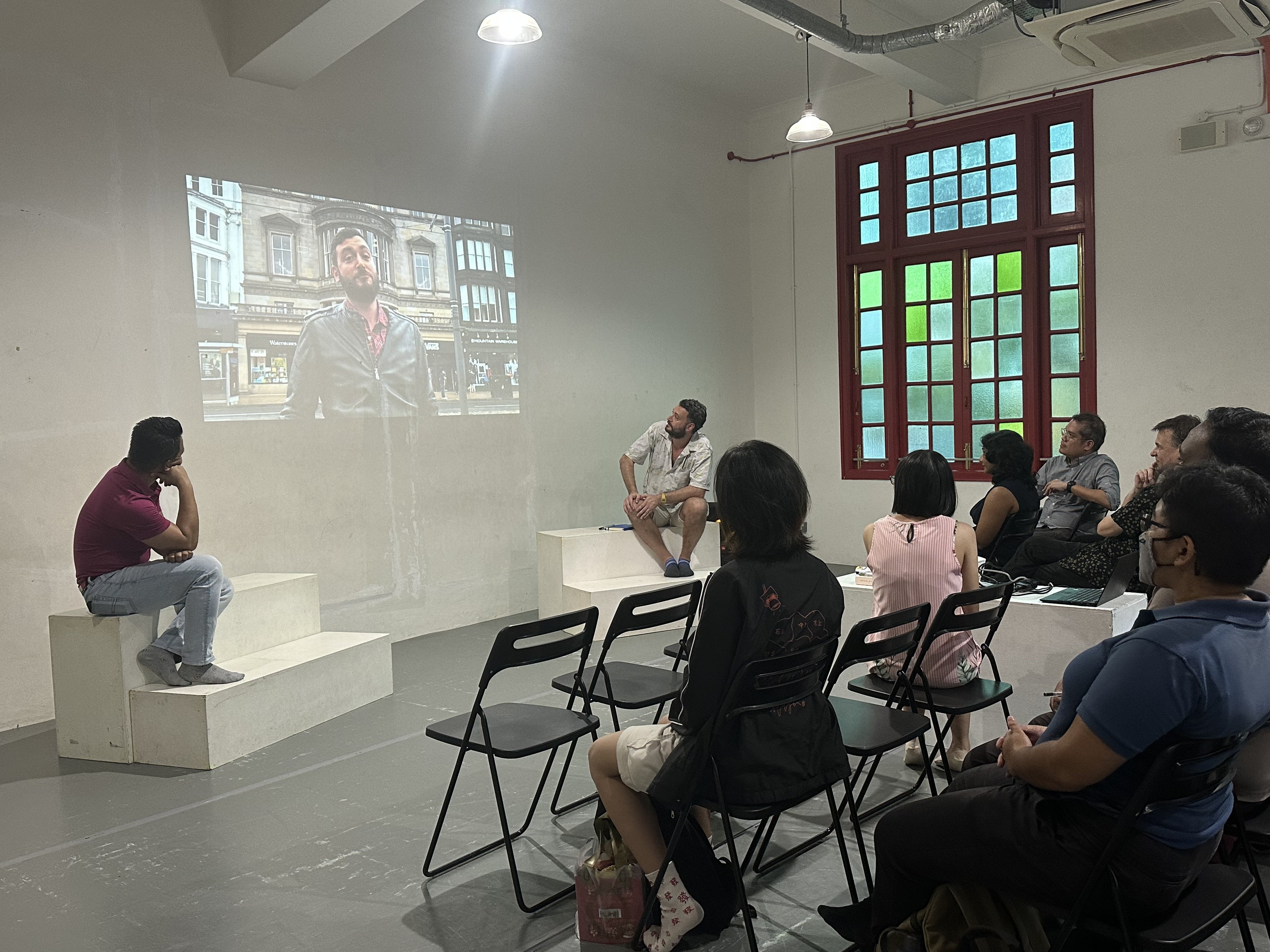 A photograph of attendees looking towards a projection on a blank wall. The projection features James standing in front of a row of shophouses.