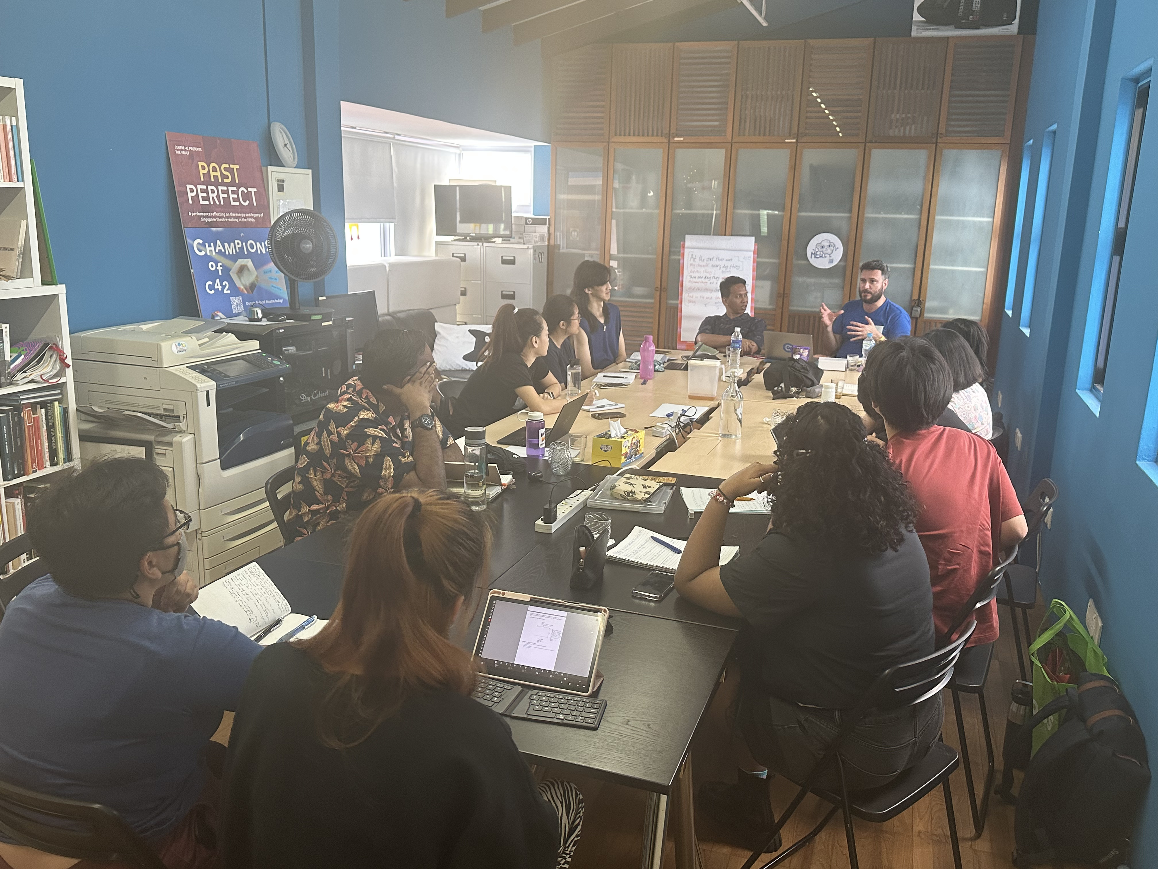 A photograph of workshop attendees gathered around the table, looking towards James and Ahmad at the head of the table.