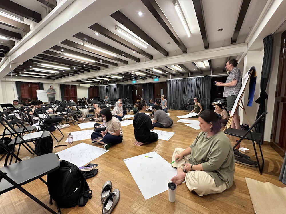 A room full of people seated on the floor with large sheets of paper, drawing and writing. A room full of people seated on the floor with large sheets of paper, drawing and writing.