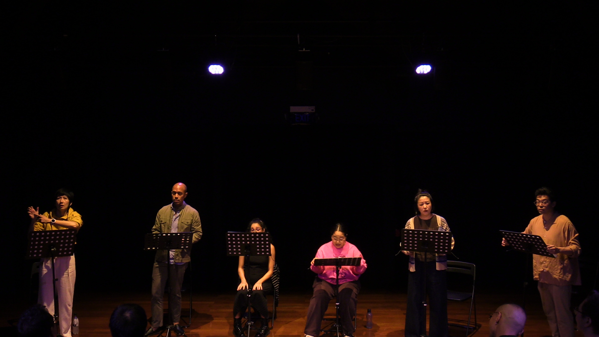 Six persons in a row of chairs, standing and sitting in front of music stands, dimly lit on a stage.