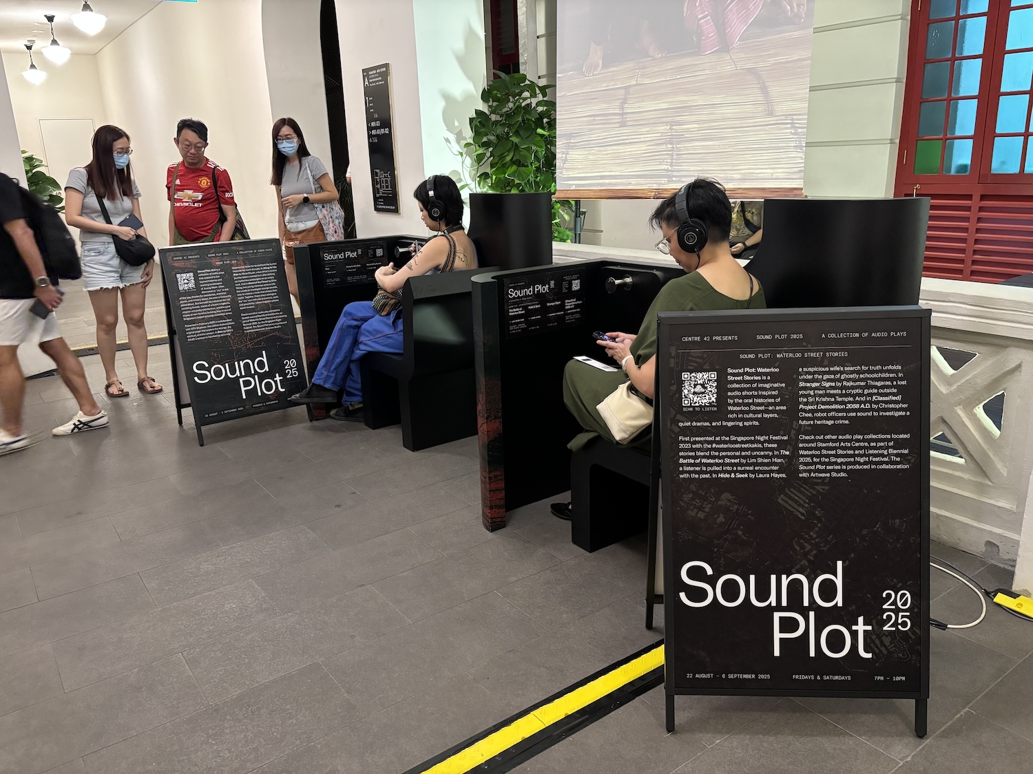 Two persons seated at small booths in a well-lit corridor, wearing headphones. Three others stand and read a signboard near them.