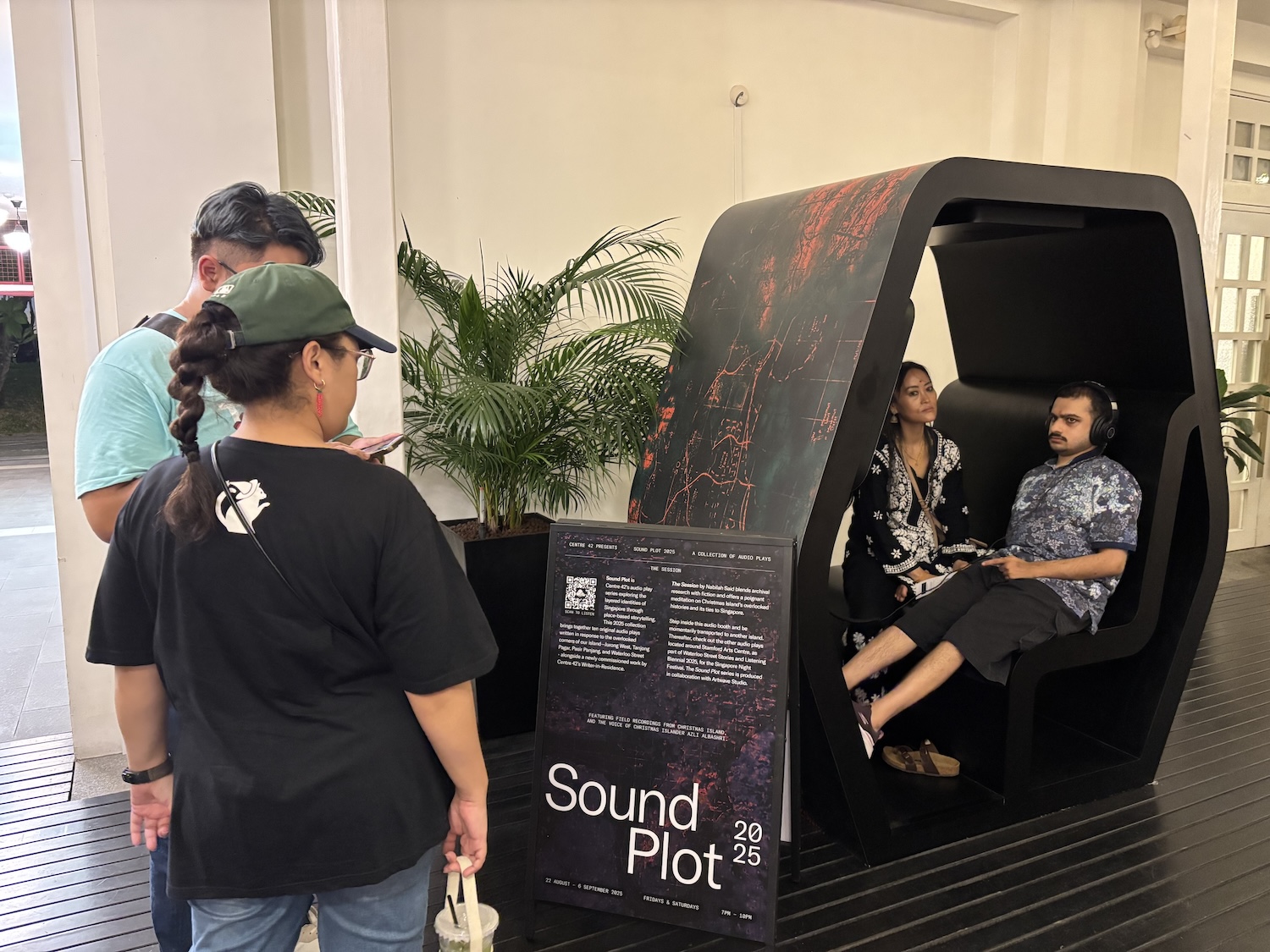 Two persons seated in a large listening booth. Another two stand in front of the booth, reading a signboard.