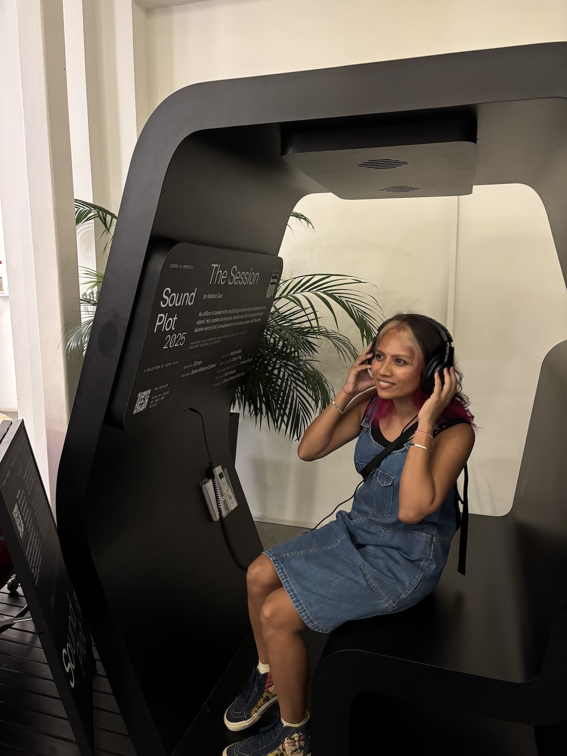 A female-presenting person seated in a large listening booth, holding headphones against her ears and smiling.
