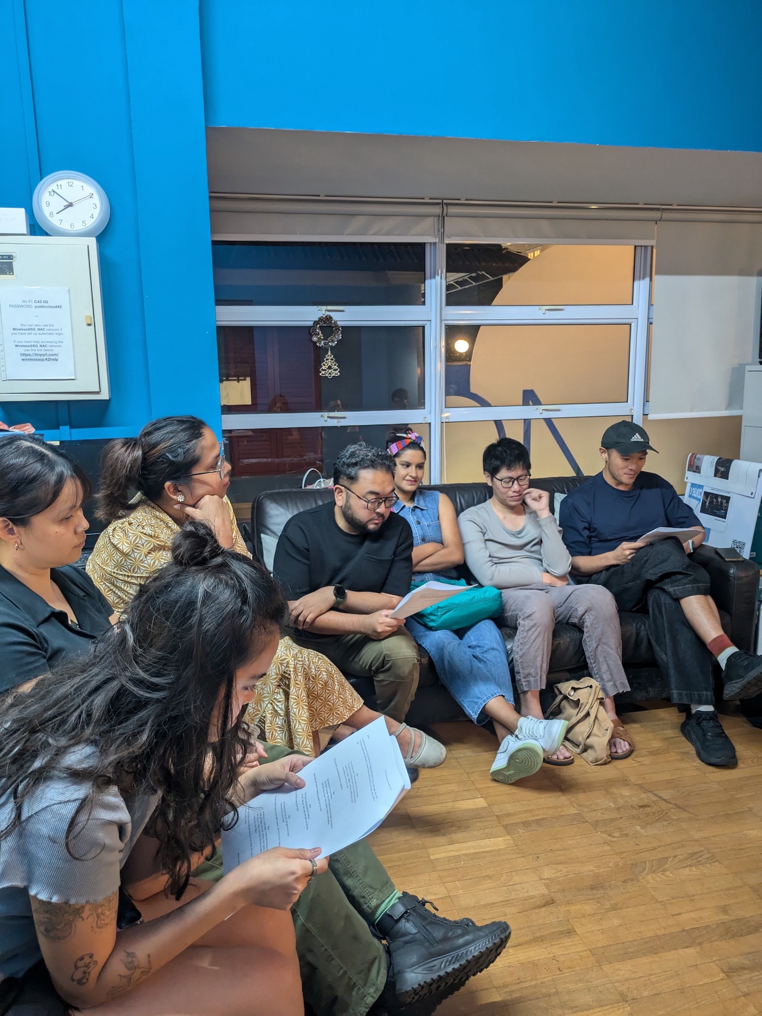 A small group of people seated on chairs and a couch in a circle. Some are reading from scripts.