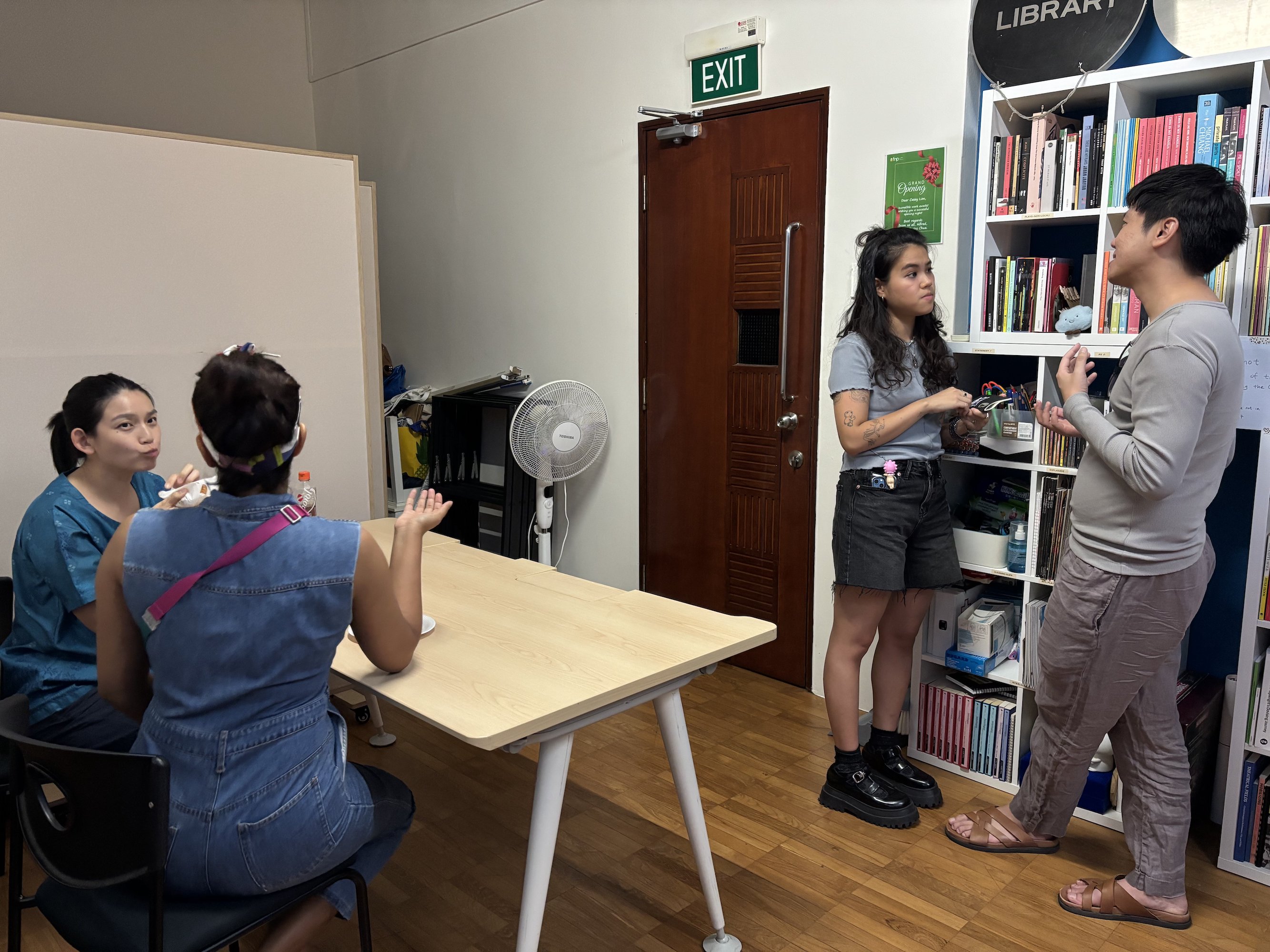 Two people seated at a table, having a conversation. Two other people stand in front of a nearby bookshelf, also conversing.