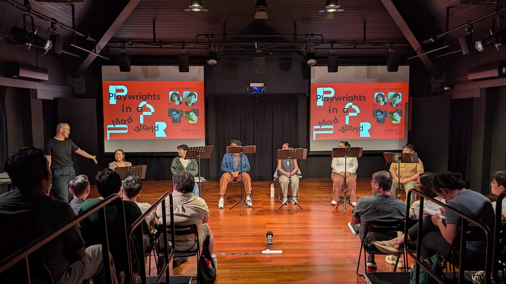 Six people seated behind music stands, in front of two projector screens. A person to their right gestures towards them.
