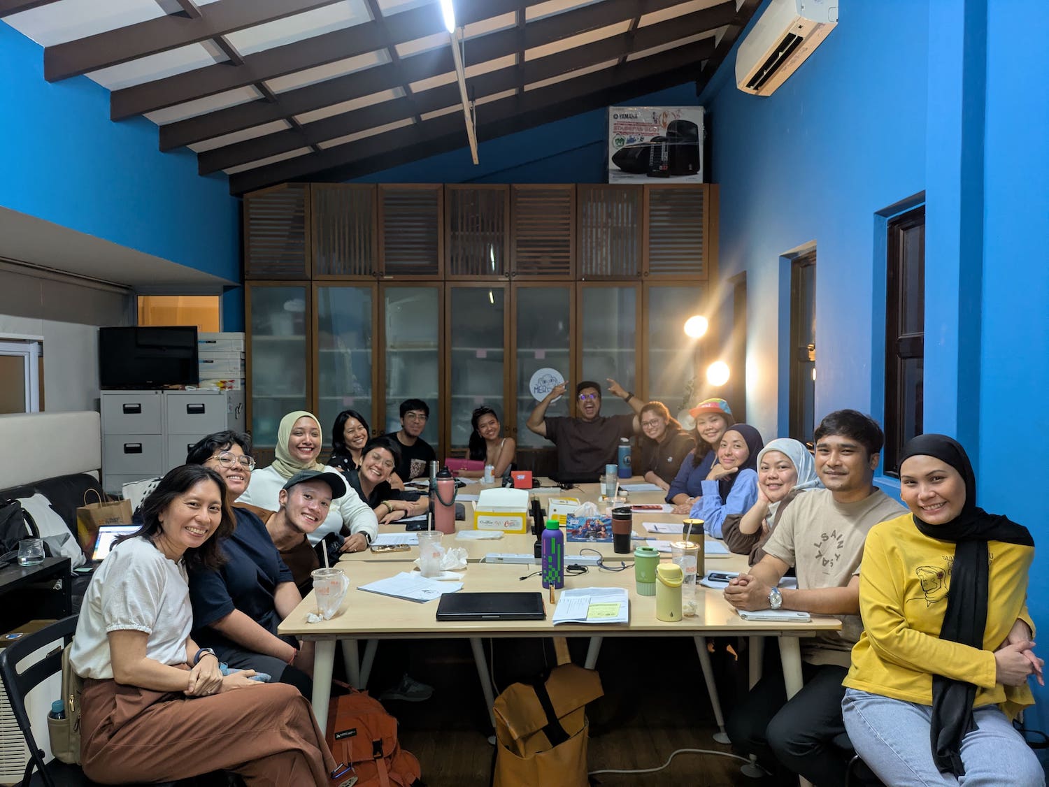 A large group of people seated around tables in an office, smiling at the camera.