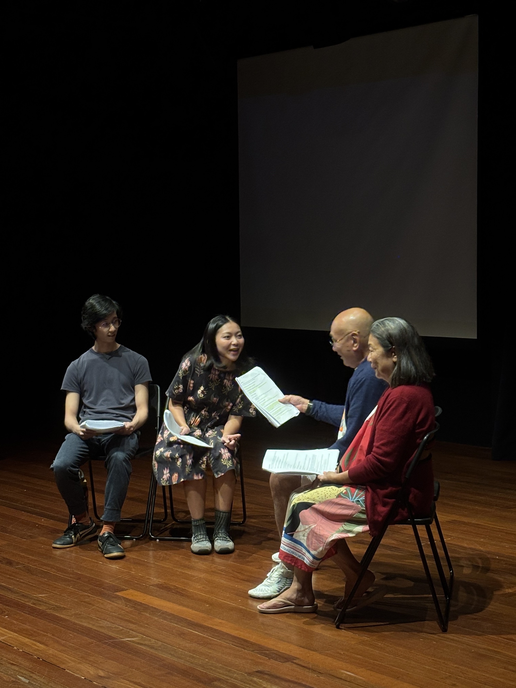 Four persons seated on chairs, facing each other and speaking. They are holding scripts.