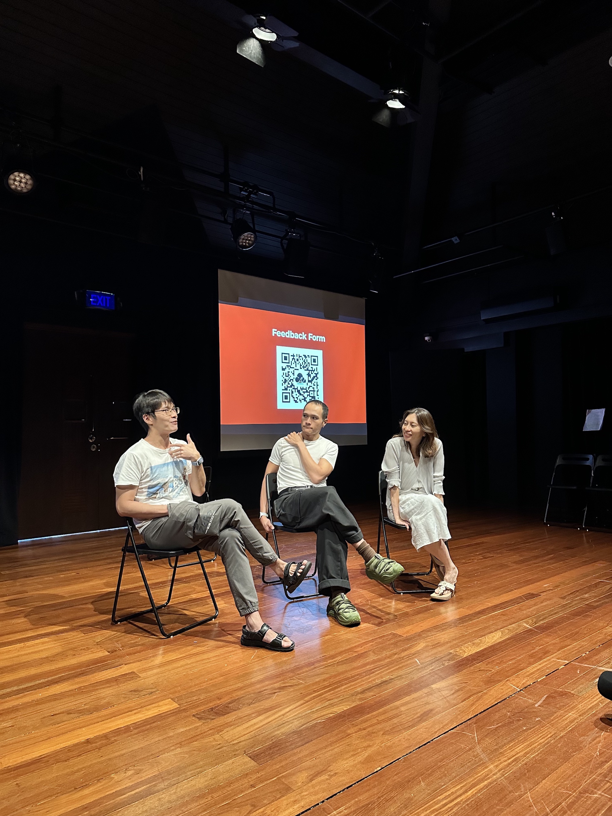 Three persons seated in a row in chairs, speaking to one another.