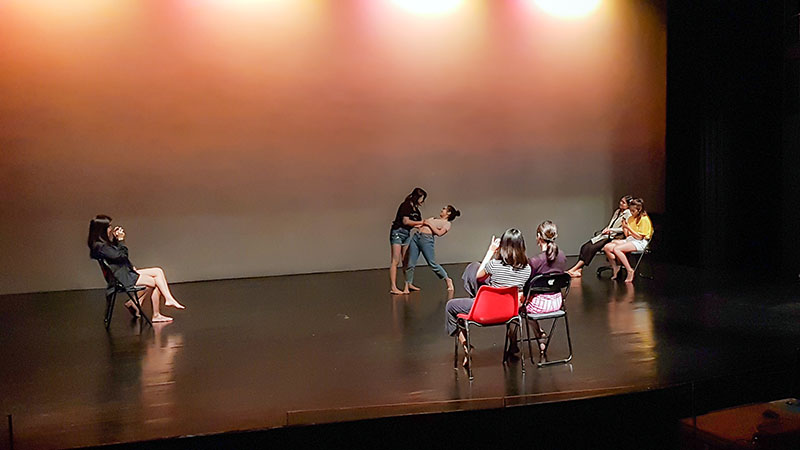 Two persons standing on a stage, one leaning backwards while the other looms over her. Around them are five persons - one seated to the left, two to the front, and two to the right - watching them.